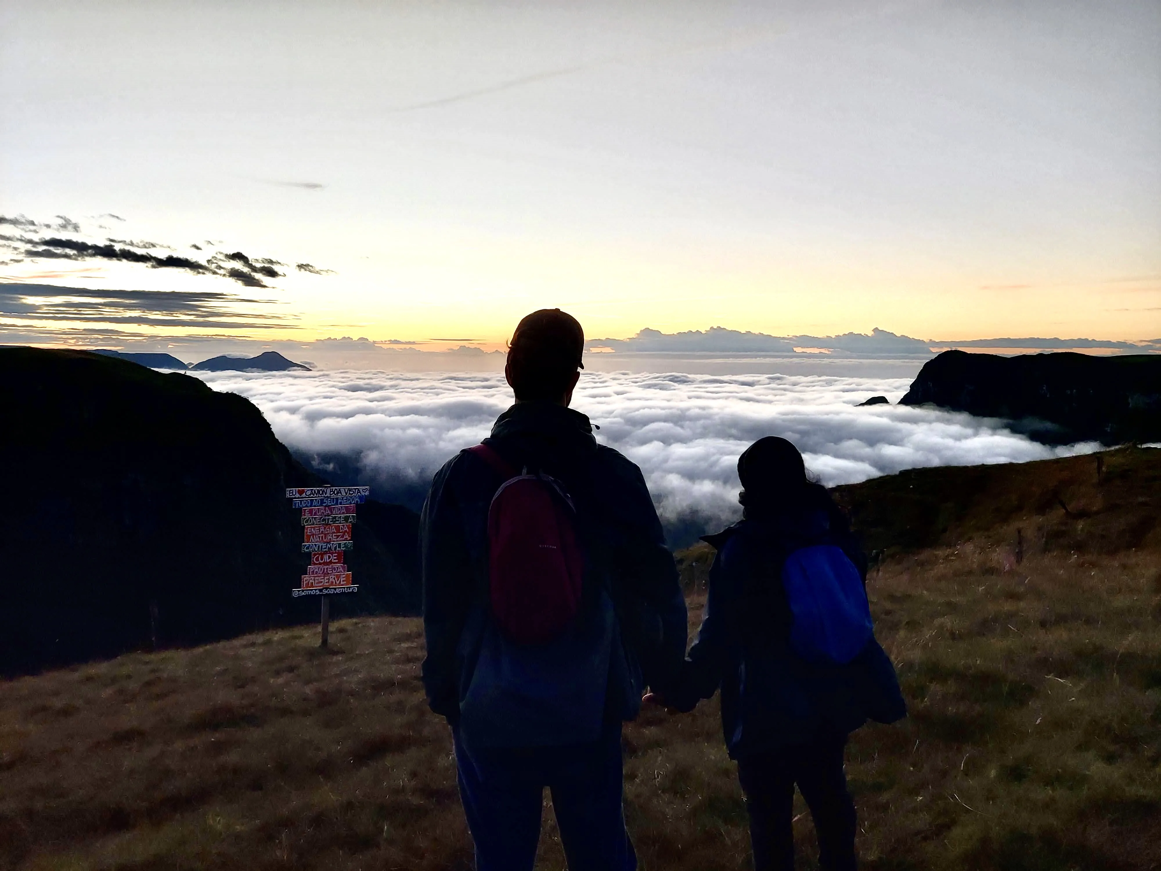 Duas pessoas vistas de costas, de mãos dadas em um campo de grama alta no alto de um cânion, observando o nascer do sol dourado sobre um imenso mar de nuvens brancas que preenche o vale entre as montanhas escuras ao fundo.