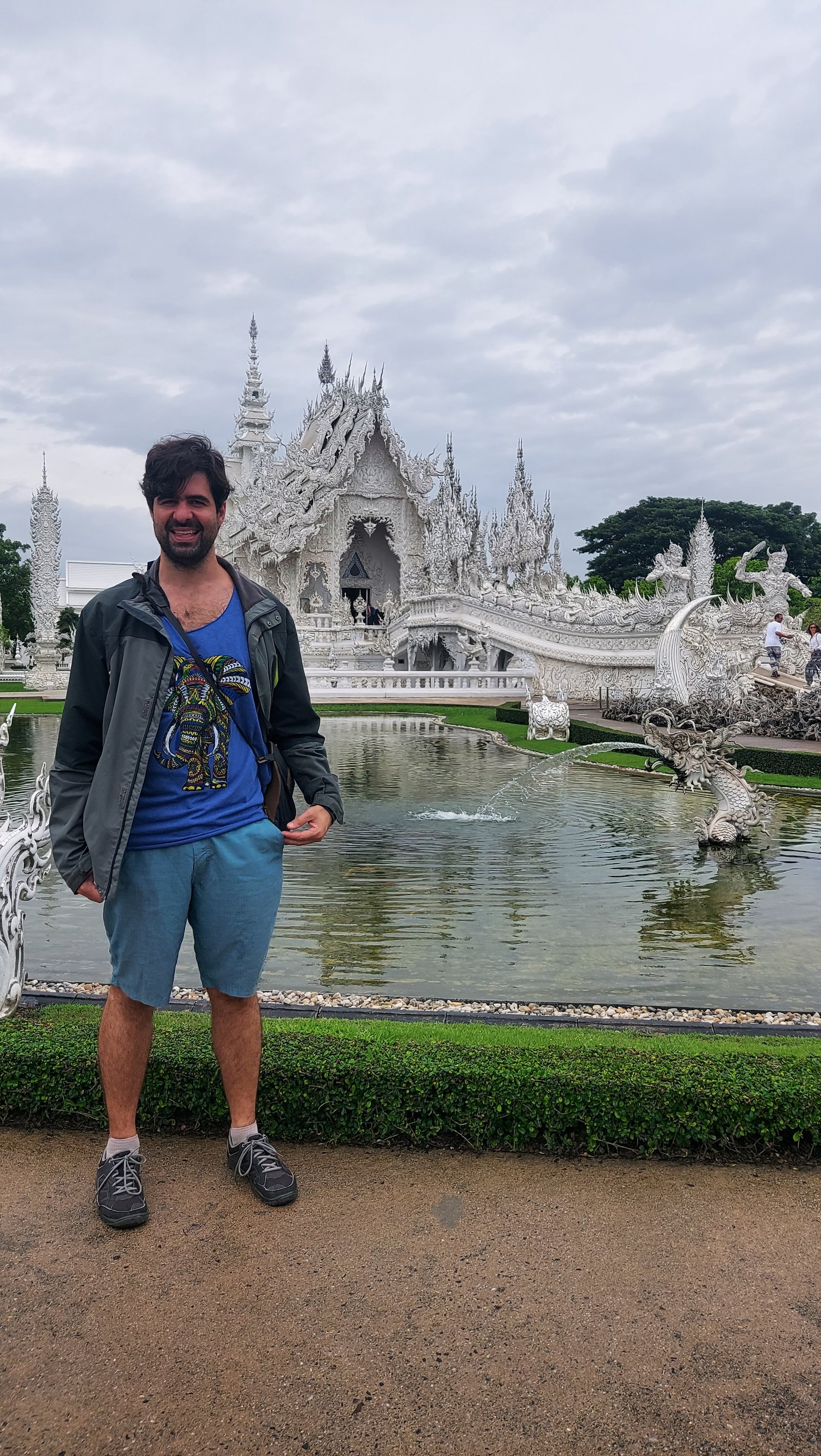 Um homem em pé à beira do lago e do jardim do Templo Branco (Wat Rong Khun) em Chiang Rai, na Tailândia, tendo a estrutura branca ornamentada ao fundo