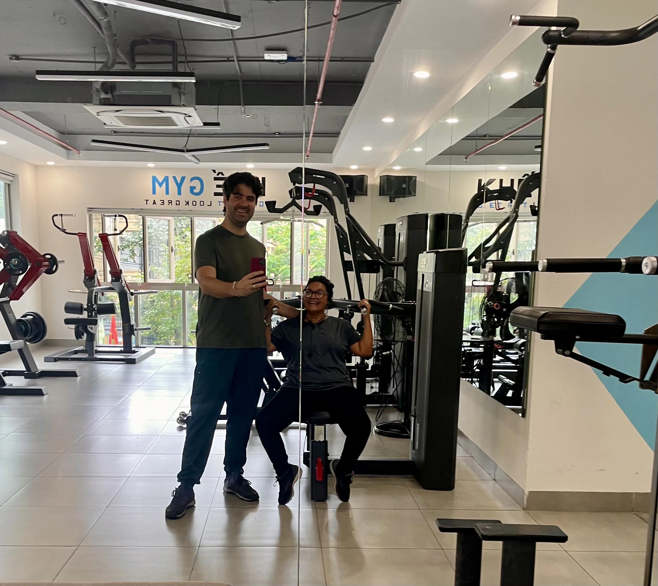Casal posando para foto no espelho de uma academia bem iluminada em Da Nang, com a pessoa em pé vestindo camiseta verde-oliva e calça azul, e a outra sentada em um aparelho de musculação de braço vestindo blusa cinza.