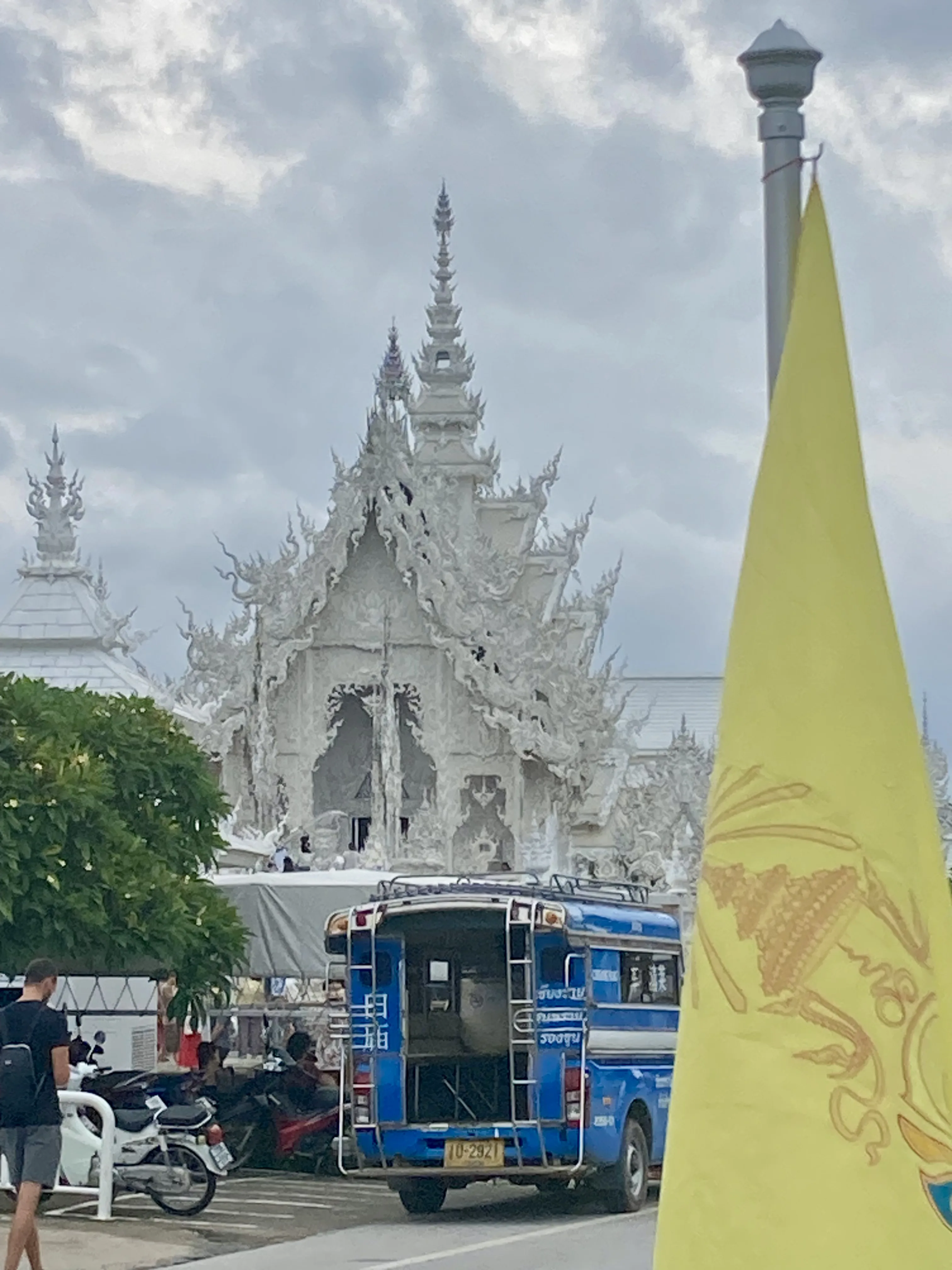 Área externa do Templo Branco em Chiang Rai, exibindo a fachada principal ricamente detalhada, uma bandeira amarela em primeiro plano e um veículo de transporte local estacionado.