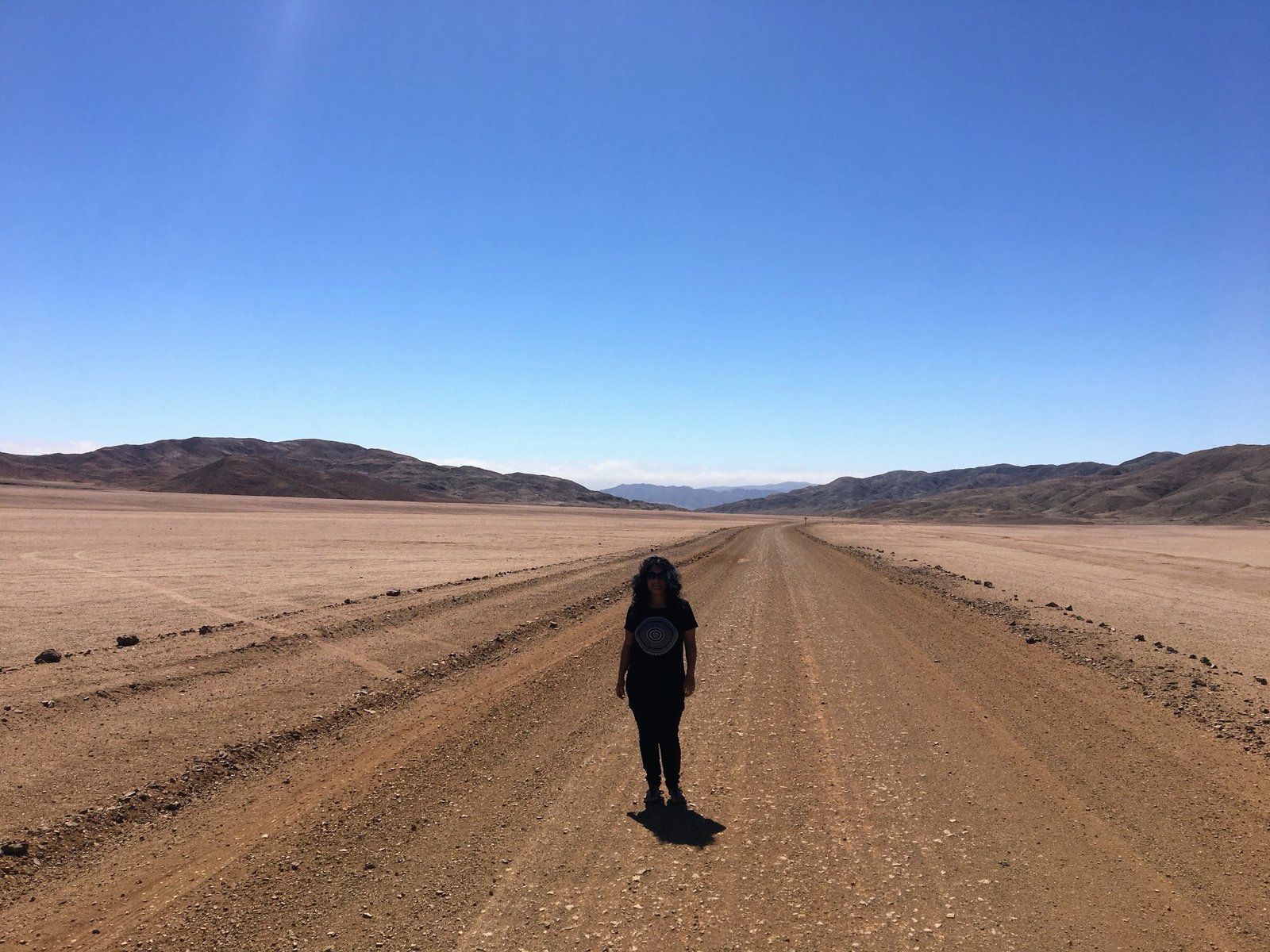 Viajante em pé, de frente, no meio de uma estrada de terra batida durante viagem de carro pelo Deserto do Atacama, ilustrando a imensidão da paisagem sob o céu azul.