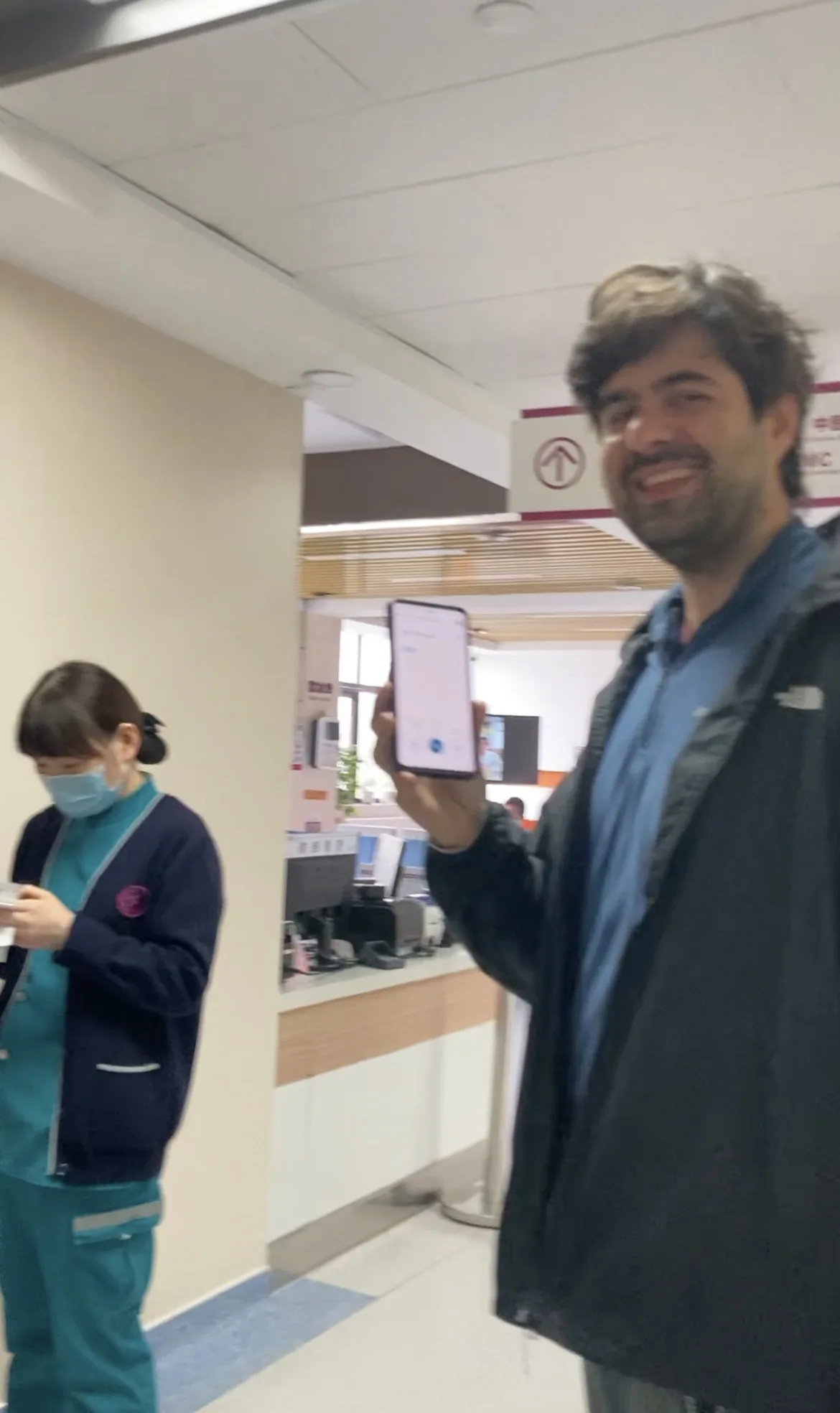 Homem sorrindo mostra a tela do celular com tradutor enquanto enfermeira de máscara anota dados na recepção de hospital na China.