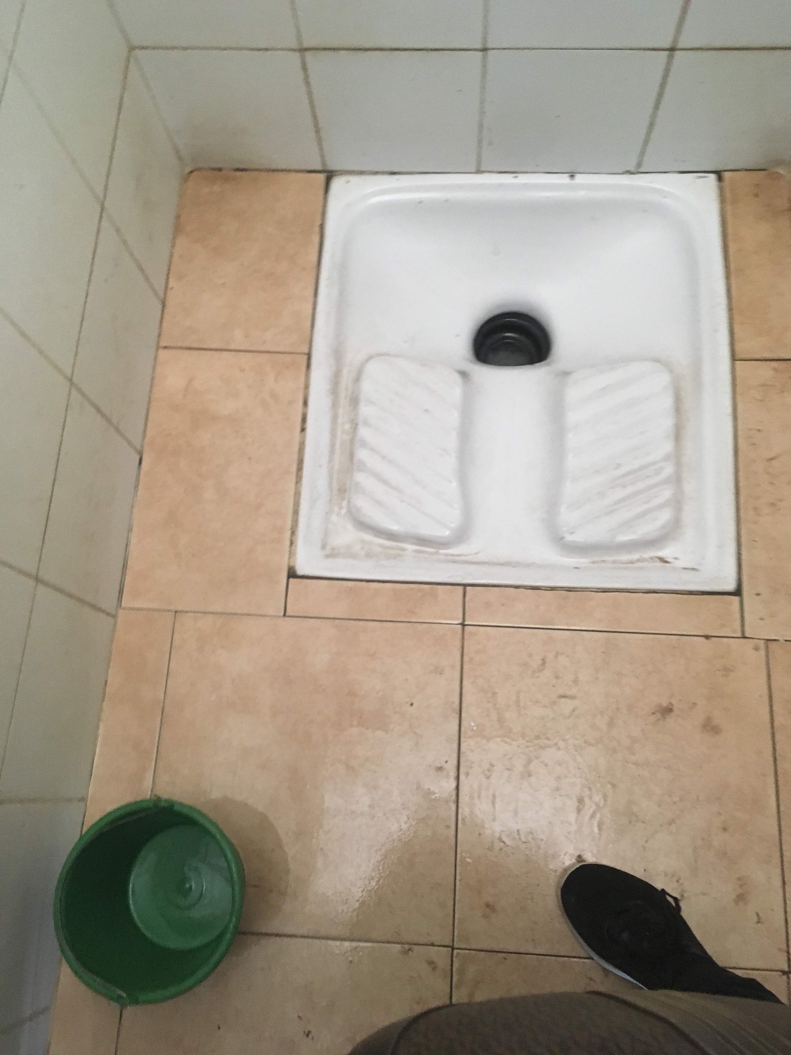 Fotografia de um banheiro turco tradicional no Marrocos mostrando uma placa de cerâmica branca instalada no chão com duas bases com ranhuras para apoiar os pés e um buraco central de escoamento ao lado de um balde verde com água
