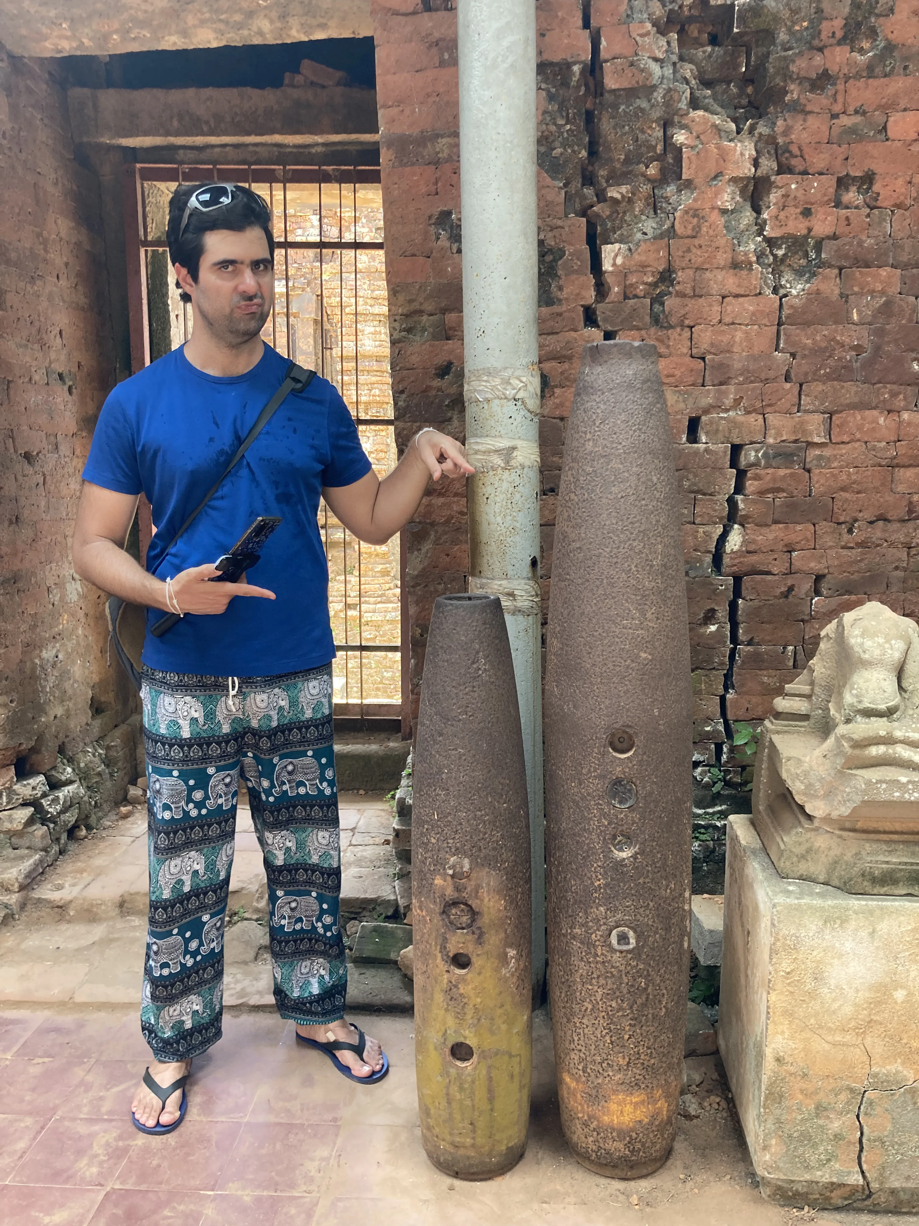 Turista a apontar para duas bombas de guerra metálicas e antigas expostas junto a uma parede de tijolos vermelhos no complexo histórico de My Son, no Vietname.