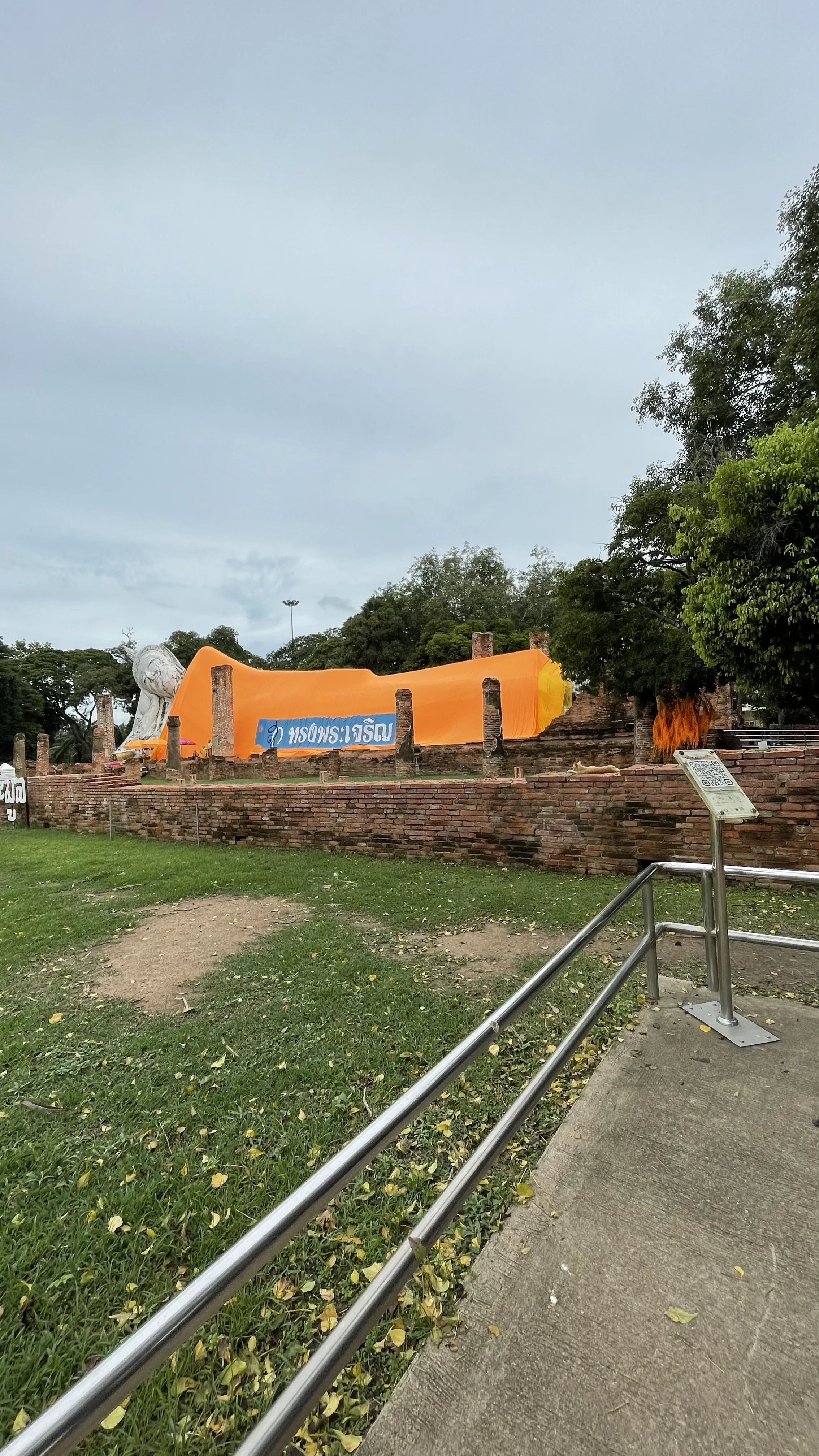 Vista ampla e ao ar livre da imensa estátua do Buda Deitado de Wat Khun Inthapramun com mais de 50 metros de comprimento, quase toda coberta por um grande manto laranja, atrás de antigas ruínas de tijolos, em um dia nublado.