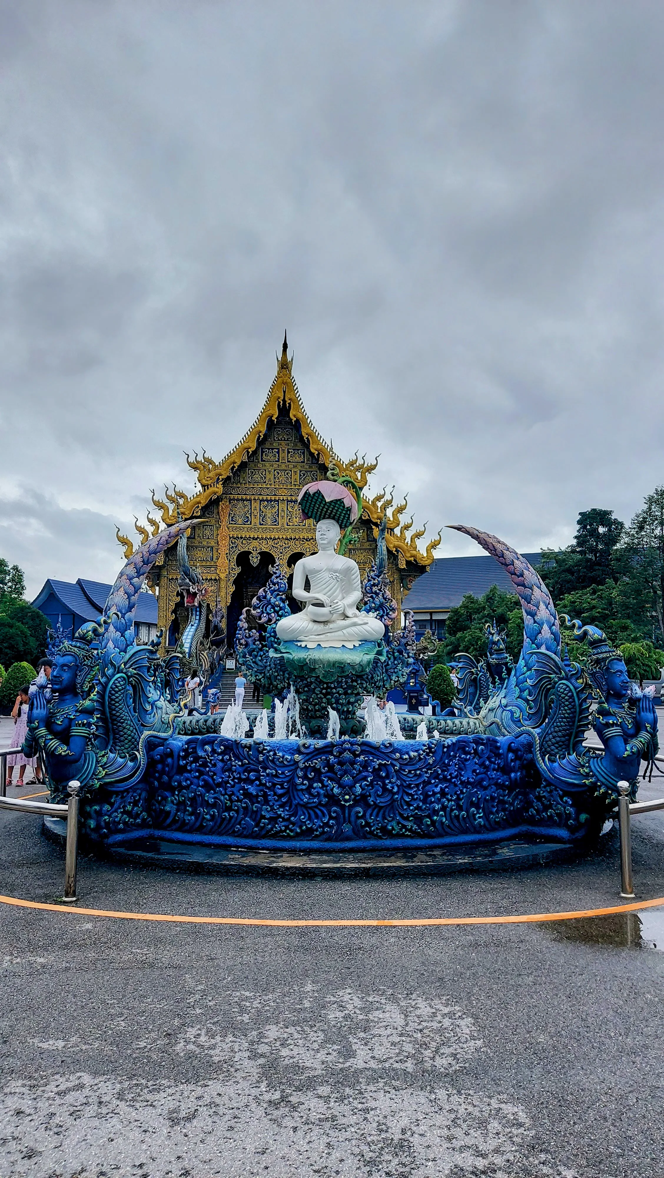 Fonte ornamental com estátua de Buda e figuras mitológicas azuis em frente à fachada dourada e azul do Templo Azul em Chiang Rai.