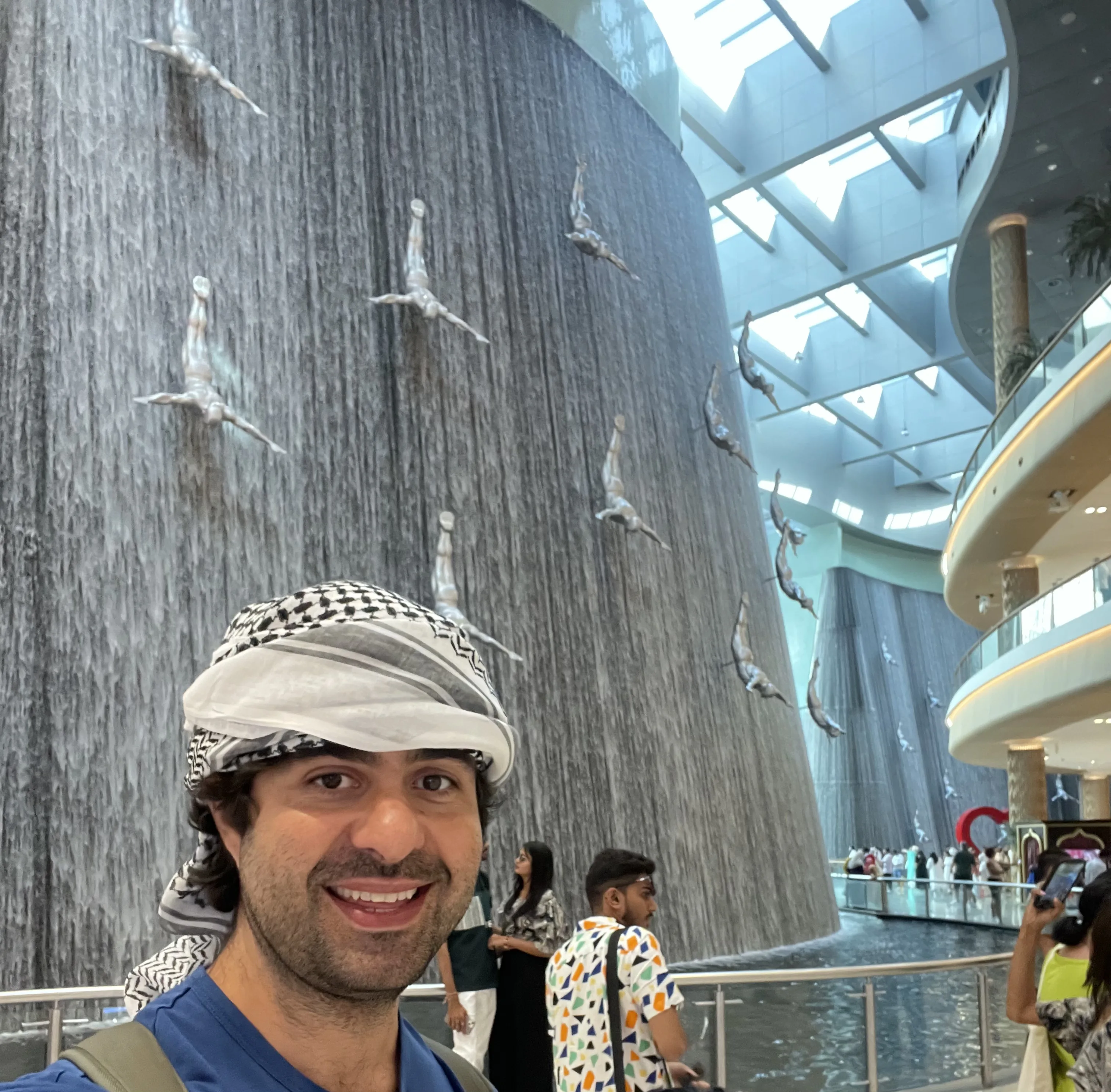 Homem em primeiro plano usando lenço xadrez keffiyeh na cabeça e sorrindo para a câmera, tendo ao fundo a imensa fonte interna em formato cilíndrico do Dubai Mall, decorada com esculturas prateadas de figuras humanas em pose de mergulho.