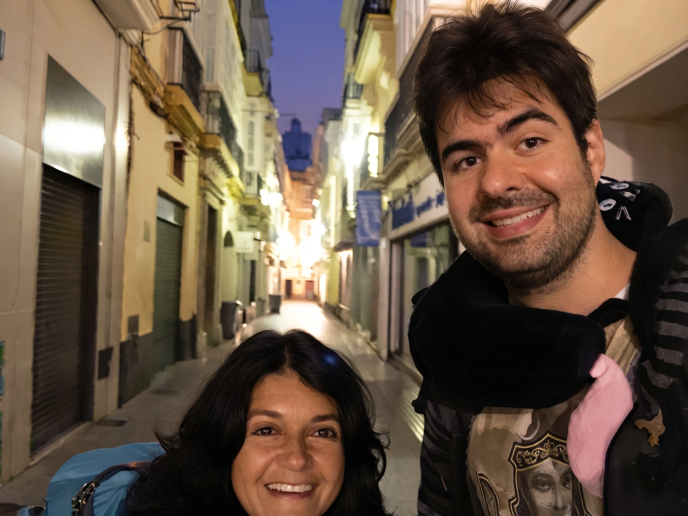 Um homem e uma mulher sorridentes em selfie durante o amanhecer em uma rua estreita e histórica ladeada por edifícios antigos em Cádiz.