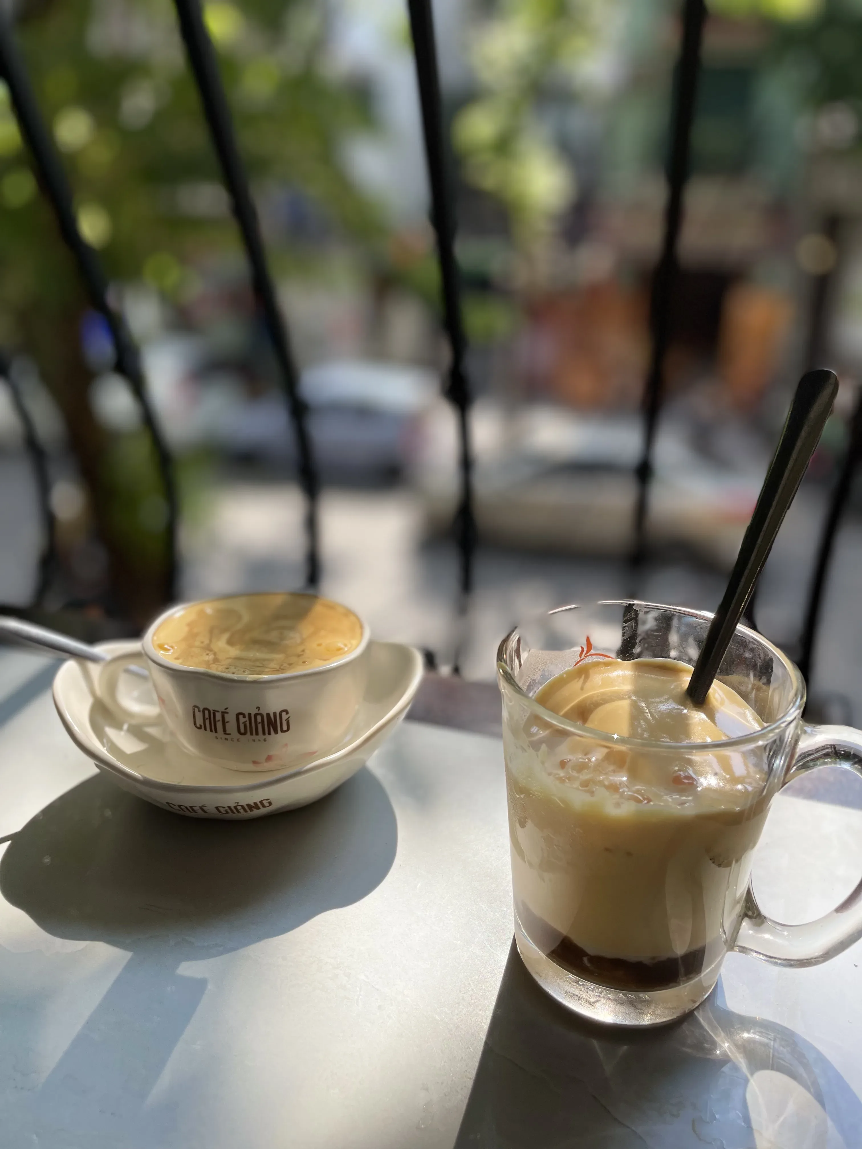 Em primeiro plano à direita, uma caneca de vidro transparente revela camadas de um café denso e escuro na base com uma espessa e cremosa cobertura amarelada de gema de ovo batida, acompanhada por uma colher preta. Ao fundo à esquerda, uma xícara de cerâmica branca com o logotipo 'Café Giang' descansa sobre um pires idêntico. A cena ocorre em uma mesa metálica clara, com a silhueta escura de grades verticais de uma janela e um ambiente externo iluminado e desfocado.