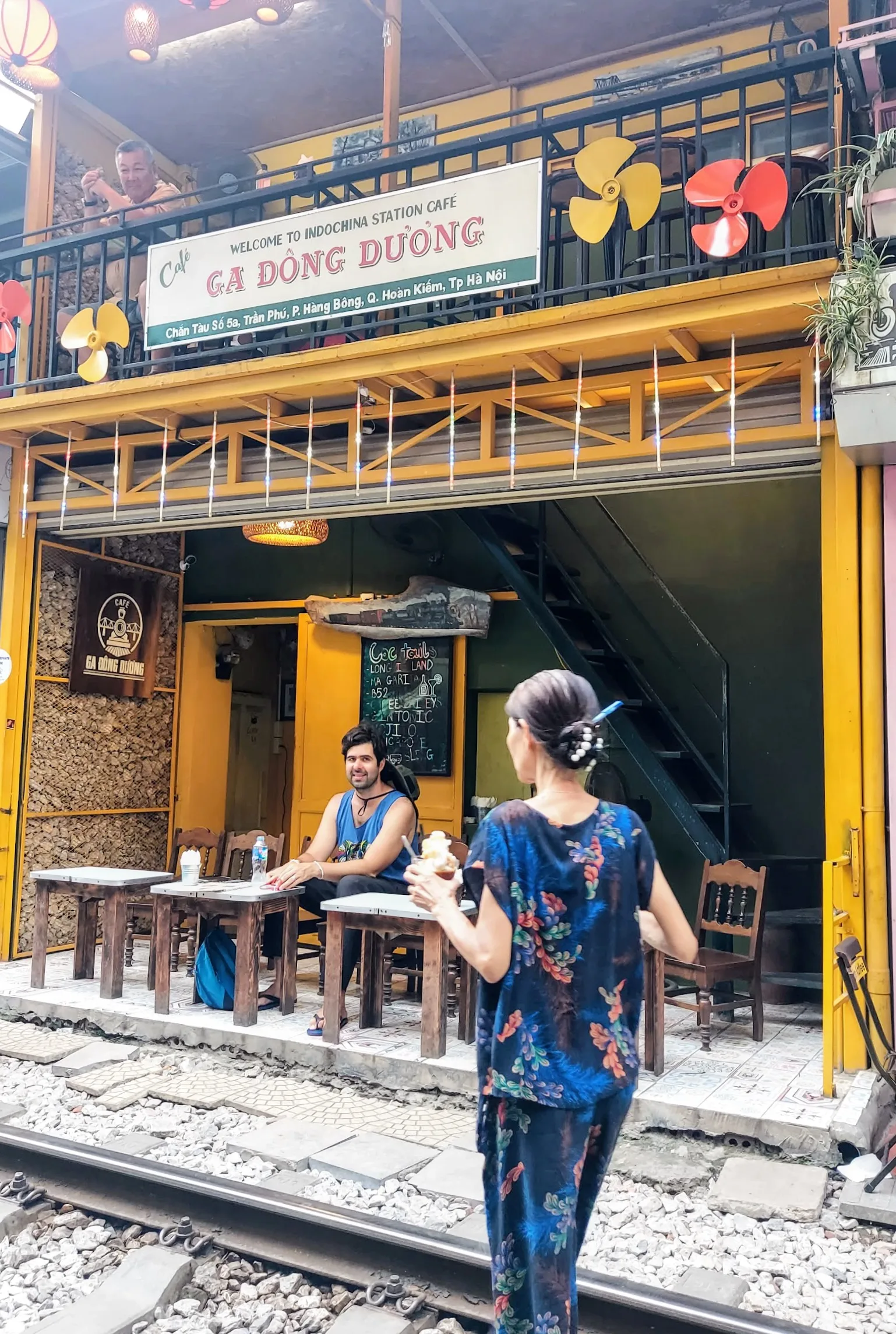 Cafeteria de fachada amarela chamada 'Ga Dong Duong' instalada à beira dos trilhos da Train Street em Hanói. Em primeiro plano, uma pessoa vista de costas e de perfil segura uma bebida, enquanto outra pessoa sentada à mesa na calçada sorri para a câmera. Ao fundo, detalhes da arquitetura de dois andares com placa informativa, ventiladores decorativos e grades pretas na sacada.