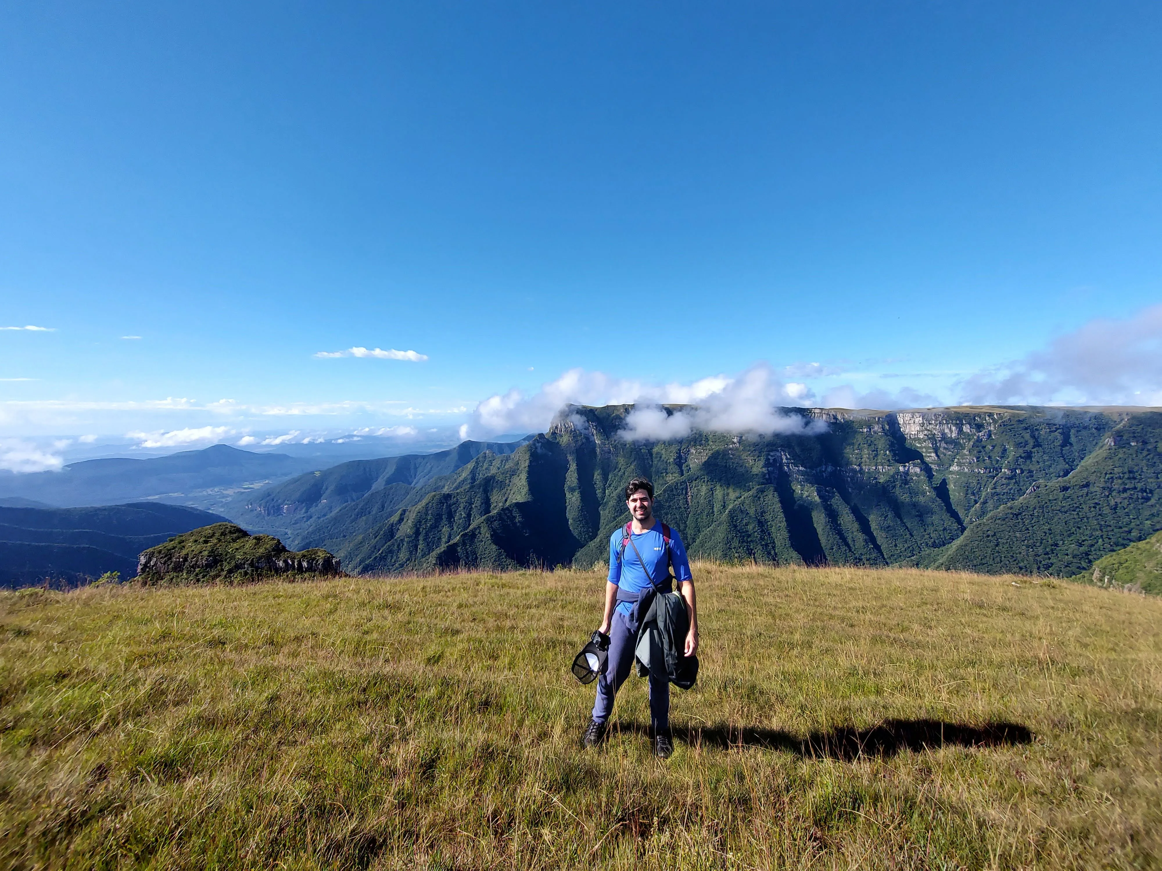 Homem em pé no alto de um campo gramado nos cânions, vestindo blusa azul e calça escura, segurando um boné e um casaco, tendo ao fundo paredões rochosos cobertos de verde e um céu azul limpo com nuvens brancas