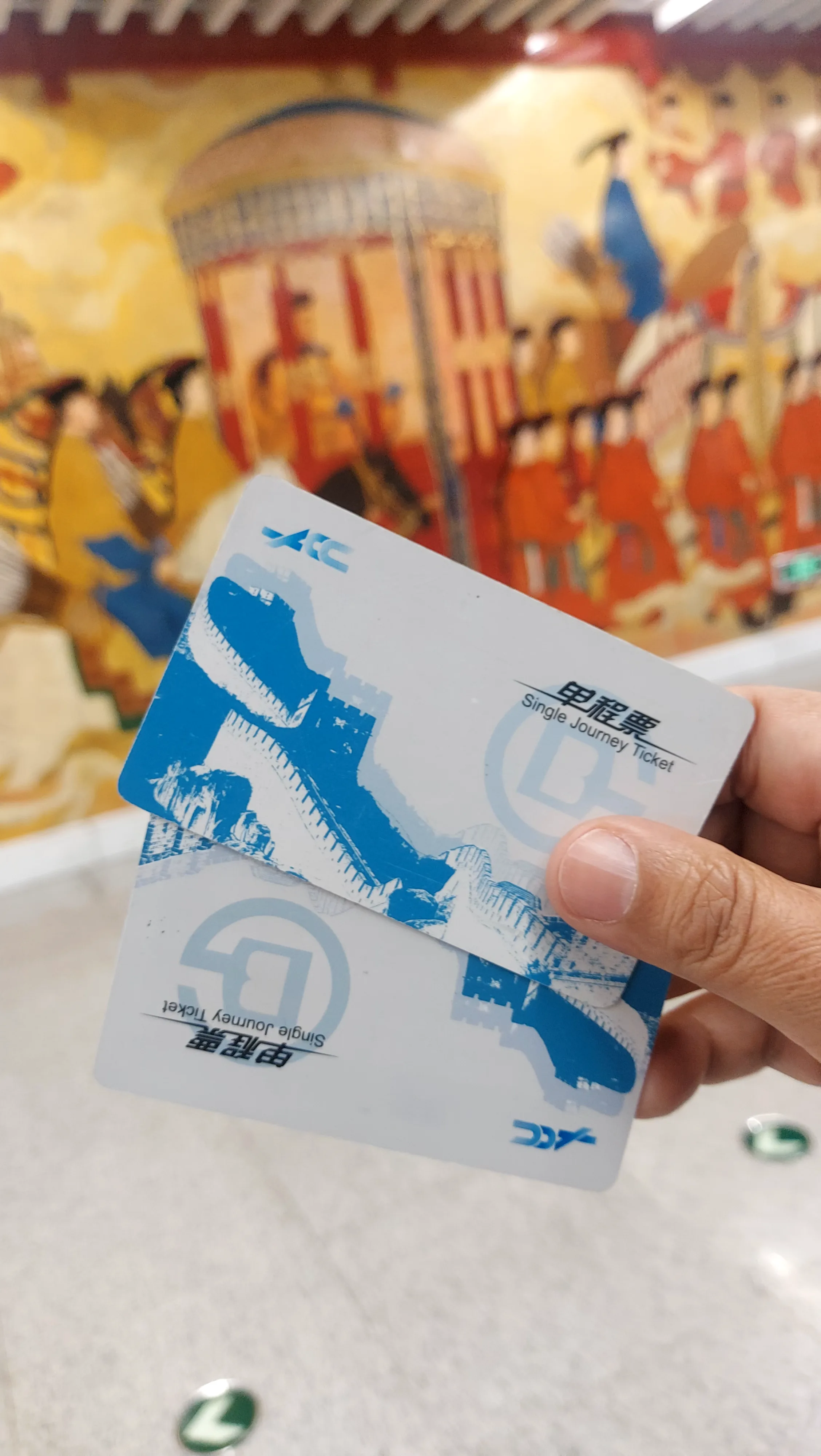 Mão segura dois cartões de viagem azul e branco do metro de Pequim com ilustração da Muralha da China diante de painel decorado.