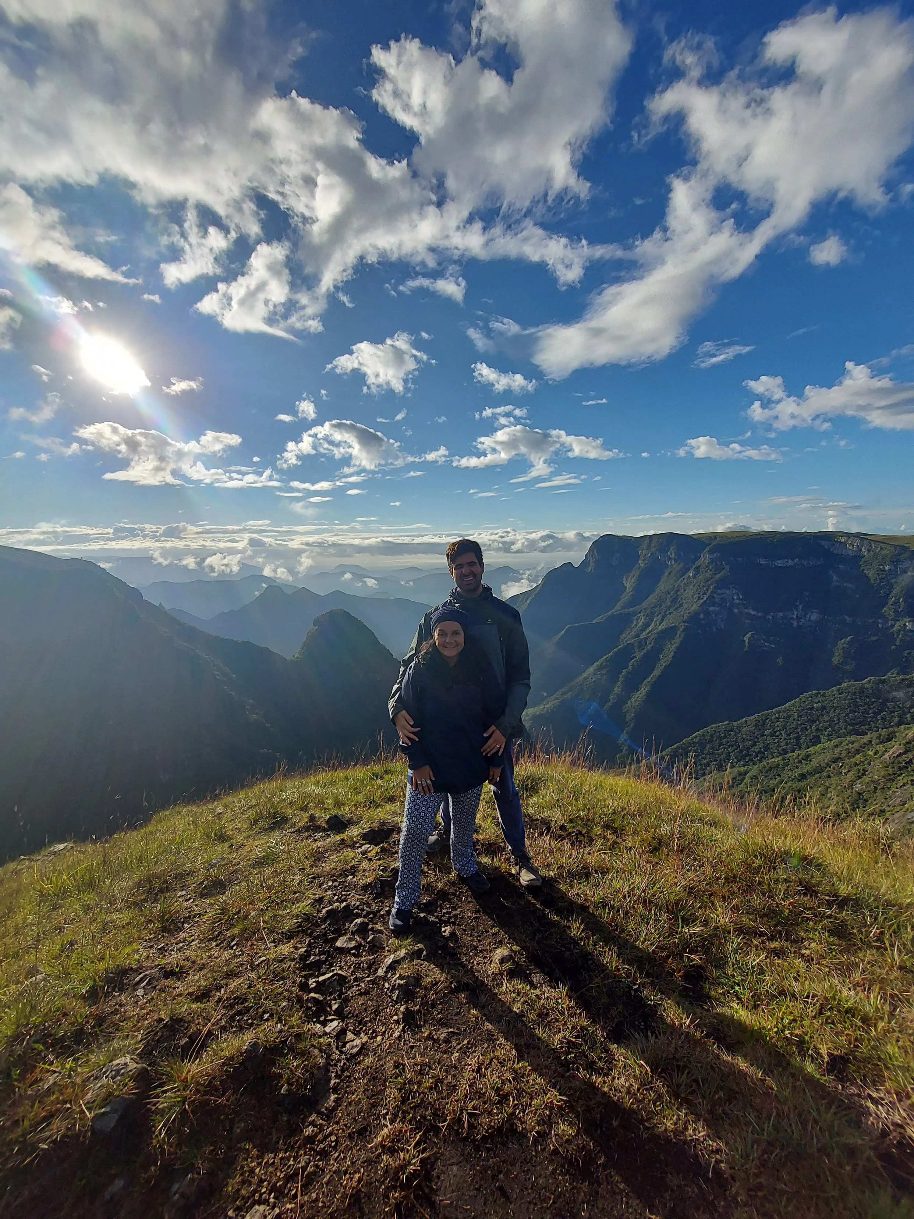 Casal posando abraçado em um cume de terra e grama nos cânions, com a luz do sol brilhando intensamente no céu azul pontilhado por nuvens brancas e montanhas cobertas de verde ao fundo.