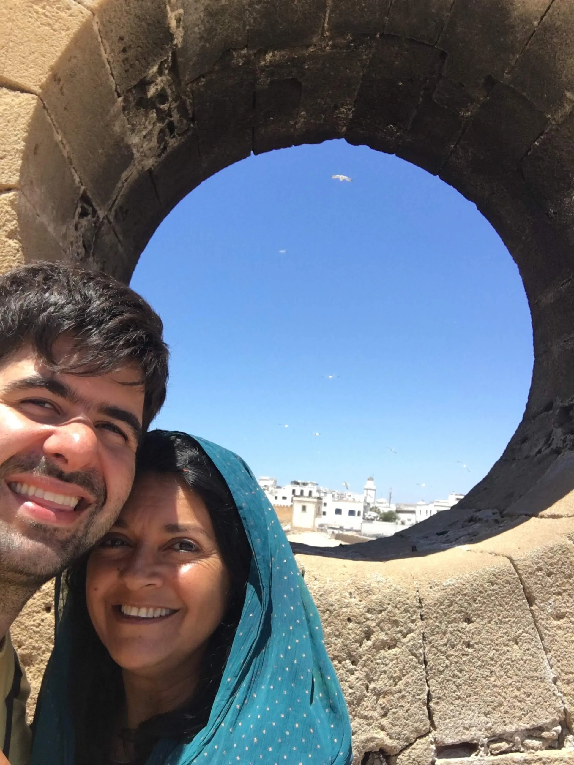 Selfie de casal sorrindo em primeiro plano enquadrado por uma grande abertura circular de pedra na muralha de Essaouira revelando o céu azul com gaivotas voando e as casas brancas da cidade ao fundo no Marrocos