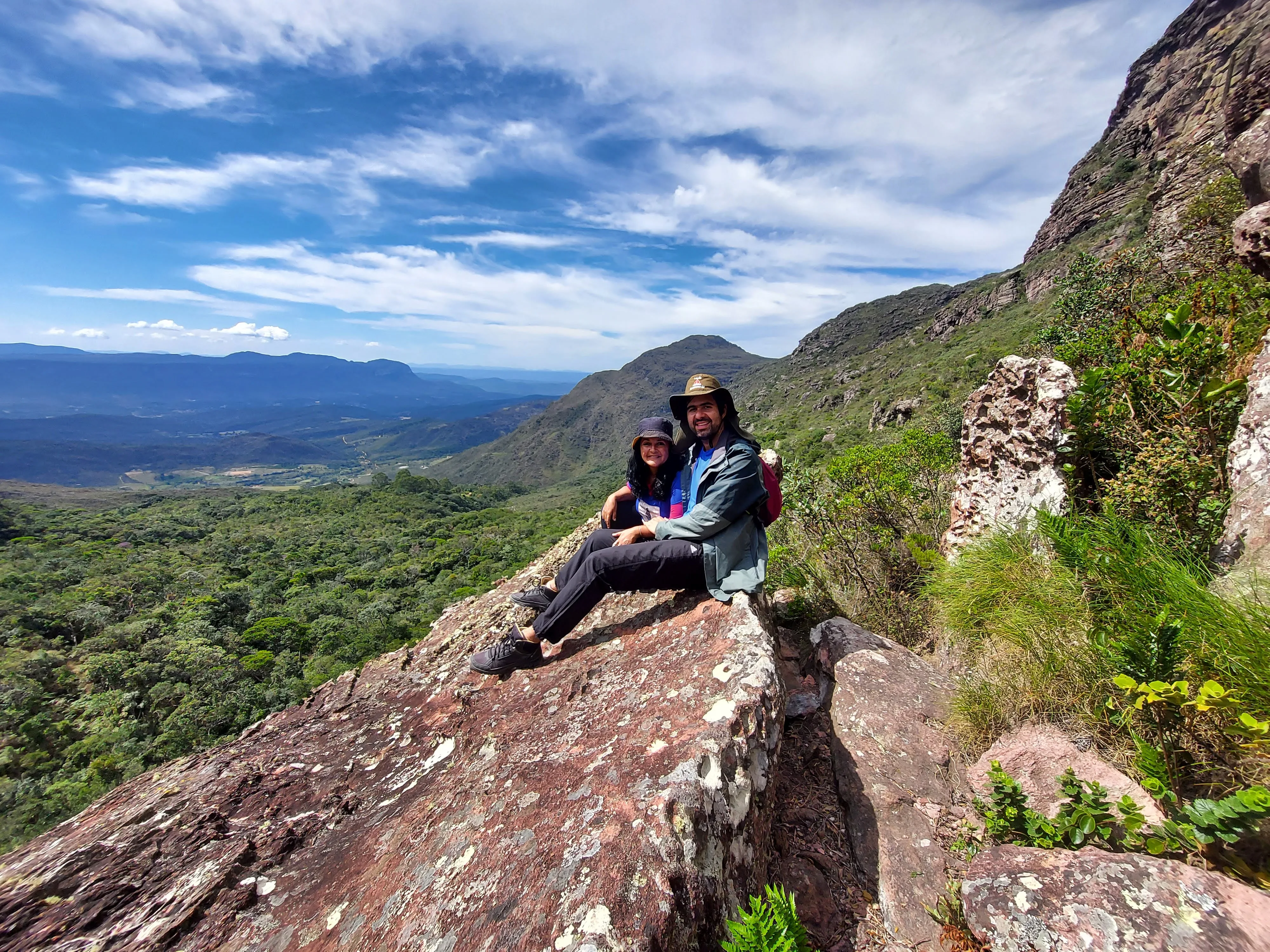 Casal feliz posa para foto sentado no topo de uma grande formação rochosa, com uma vista panorâmica espetacular de montanhas verdes e vale ao fundo sob céu azul com nuvens em dia de trekking.