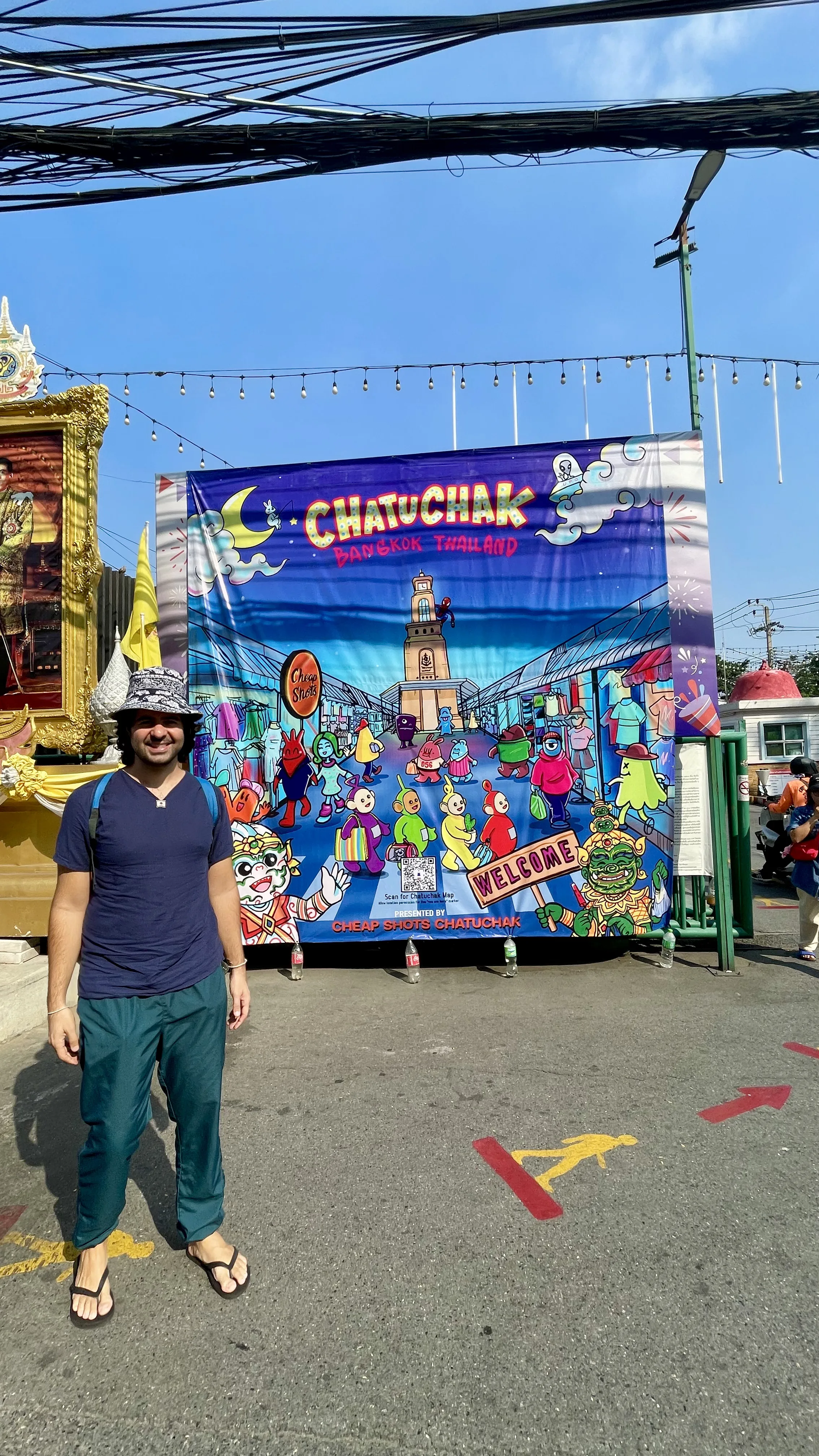 Homem em pé diante de um painel colorido de boas-vindas do Mercado de Chatuchak, em Banguecoque.