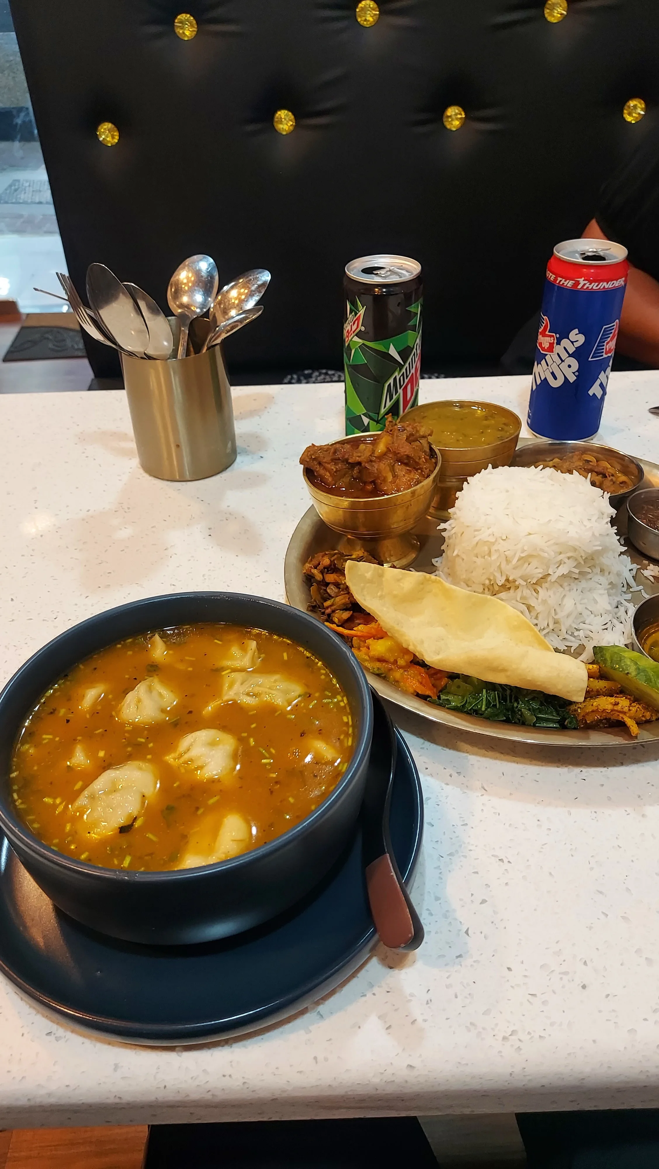 Mesa branca de restaurante exibindo um prato típico nepaleso composto por arroz branco, molhos, carnes, papadum e acompanhamentos, além de uma tigela escura com momos (bolinhos) imersos em caldo temperado, latas de refrigerante Mountain Dew e Thums Up, e talheres em um copo metálico.