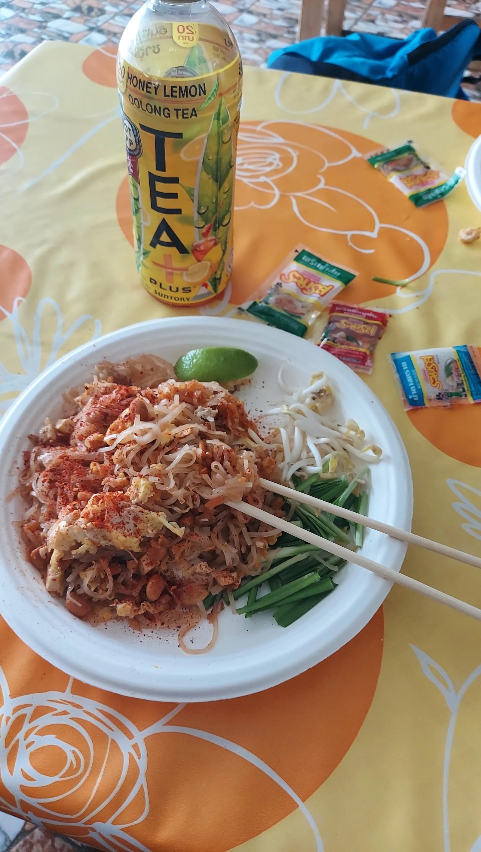 Prato de Pad Thai tailandês acompanhado de brotos de feijão, cebolinha, limão e uma lata de chá oolong com limão e mel sobre mesa colorida.
