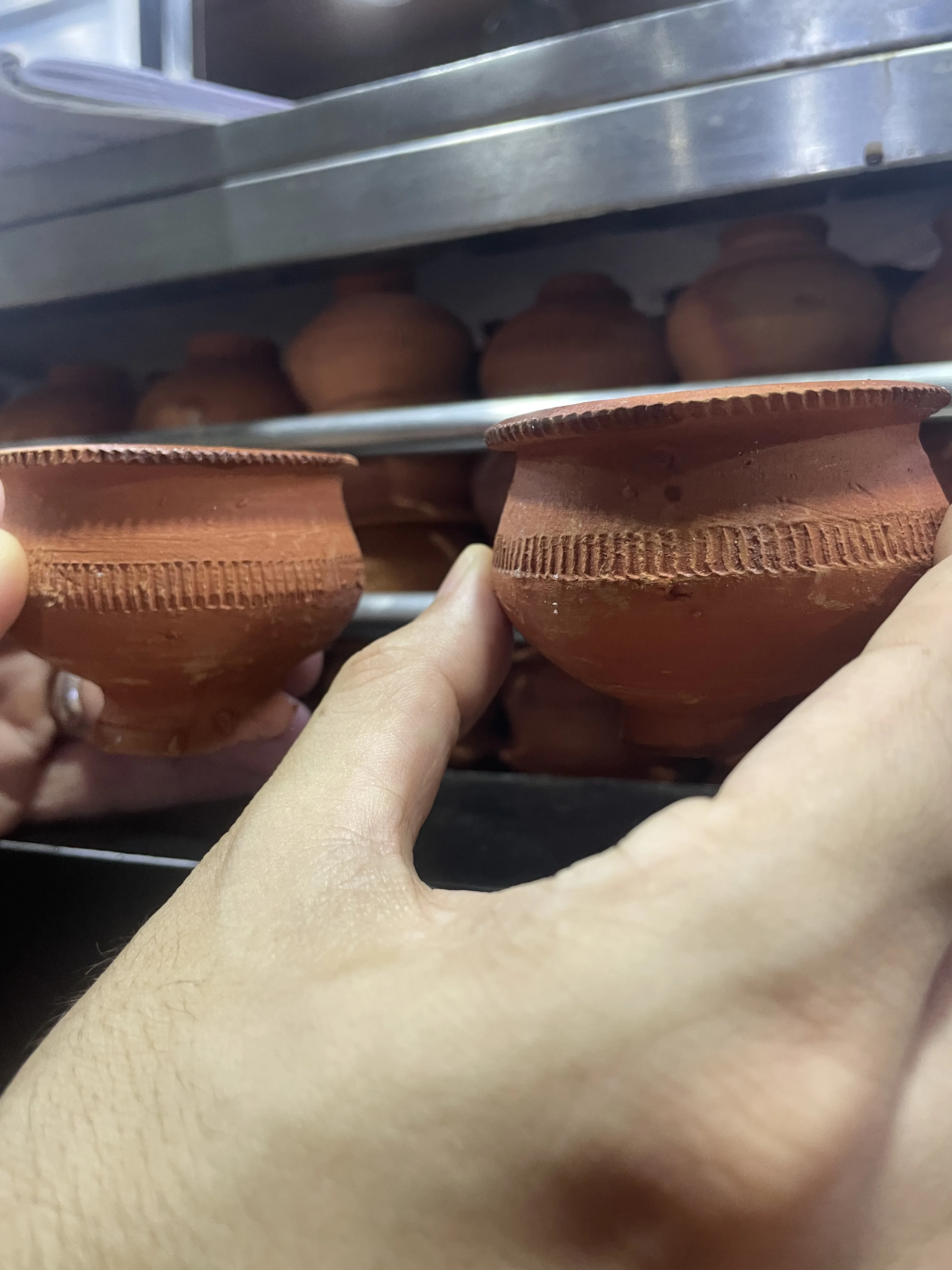 Mão segurando um pequeno copo de terracota tradicional indiano chamado Bhar, com ranhuras decorativas na borda, tendo ao fundo prateleiras metálicas com vários outros recipientes de barro.