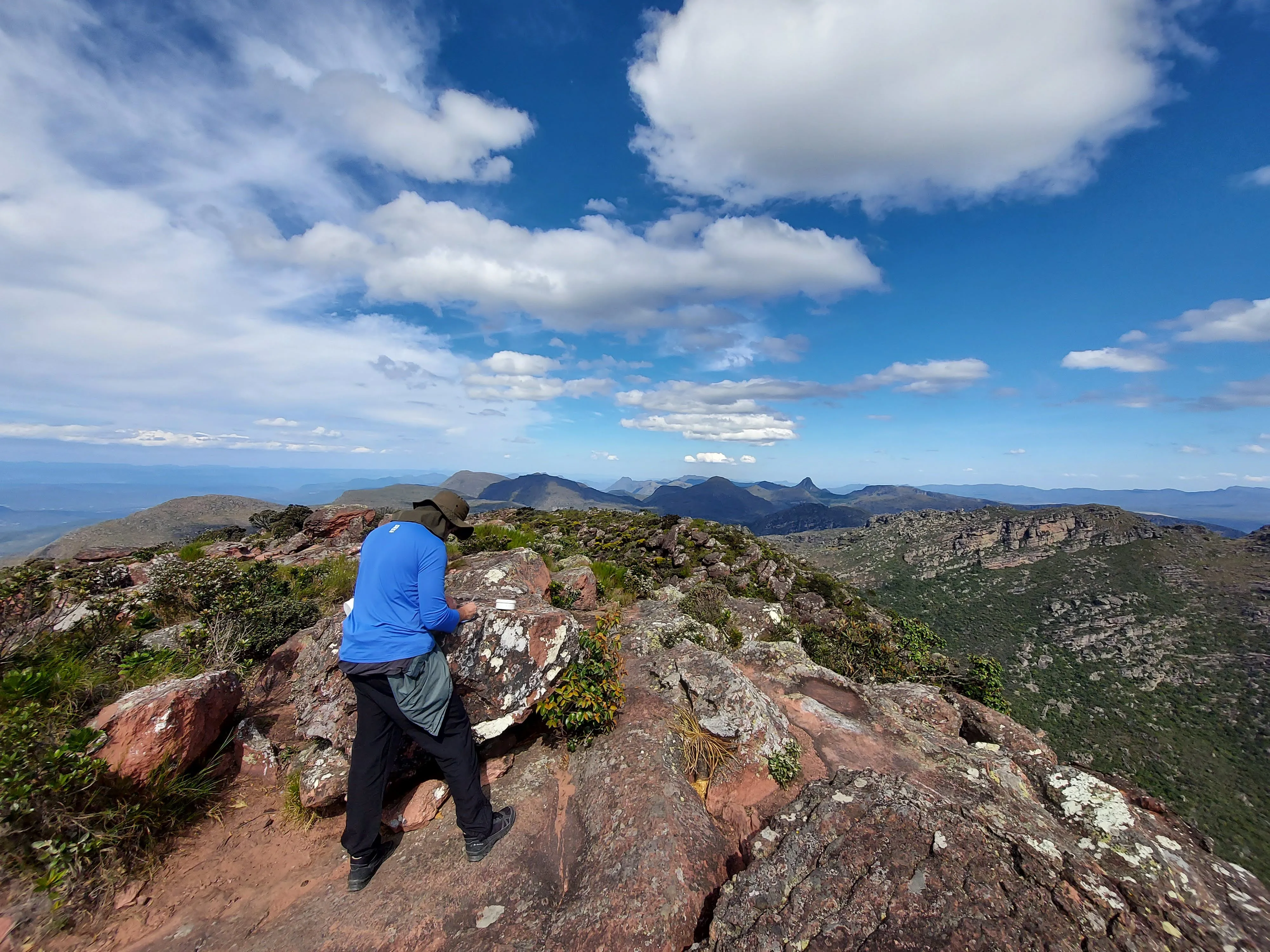 Trekking na montanha: homem usando chapéu e camisa azul anota algo em um caderno enquanto está em pé sobre rochas escarpadas, com a paisagem montanhosa e o céu nublado ao longe.