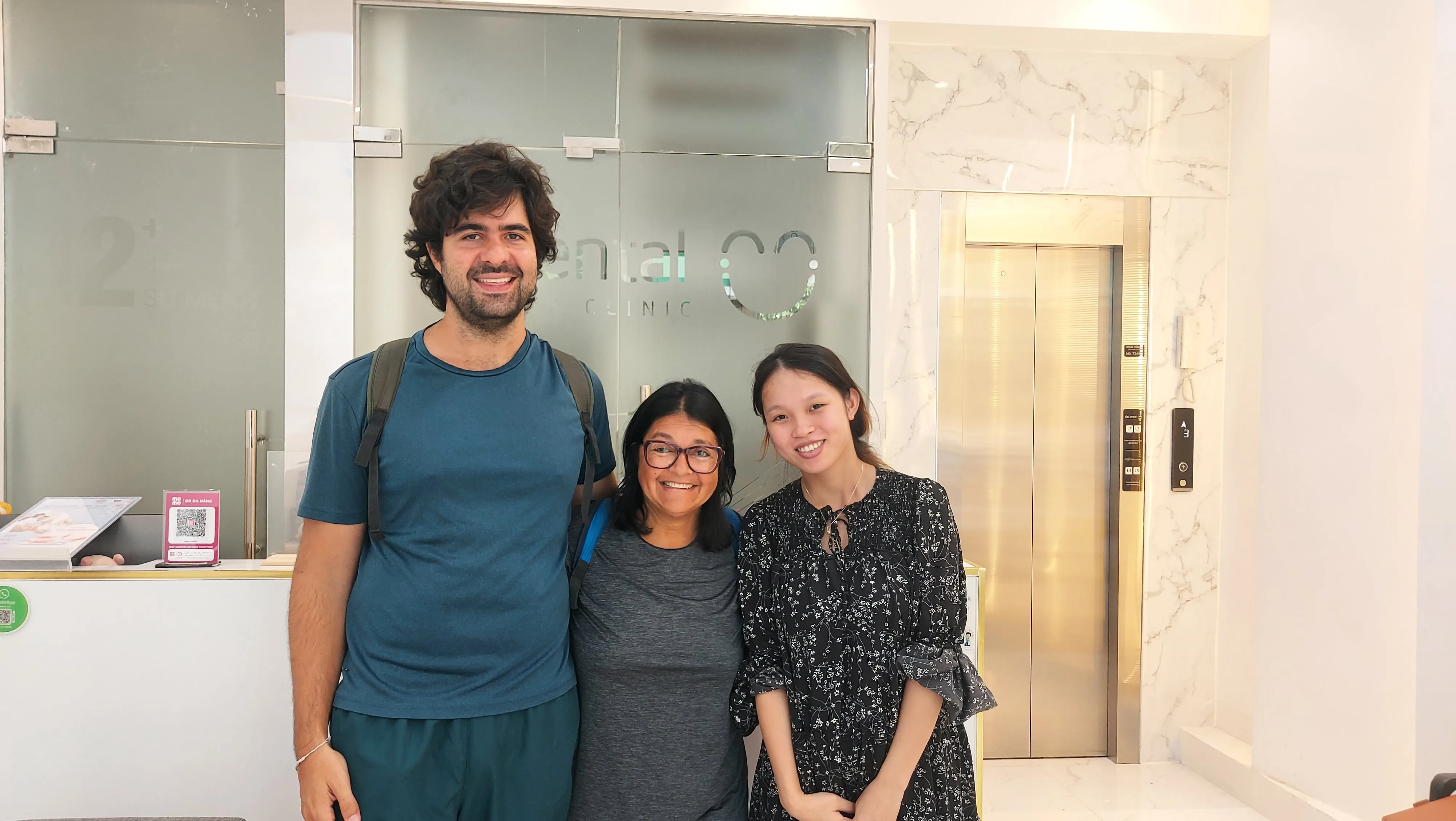 Casal de viajantes brasileiros sorrindo ao lado da atendente na recepção da clínica odontológica BeDental no Vietnã.