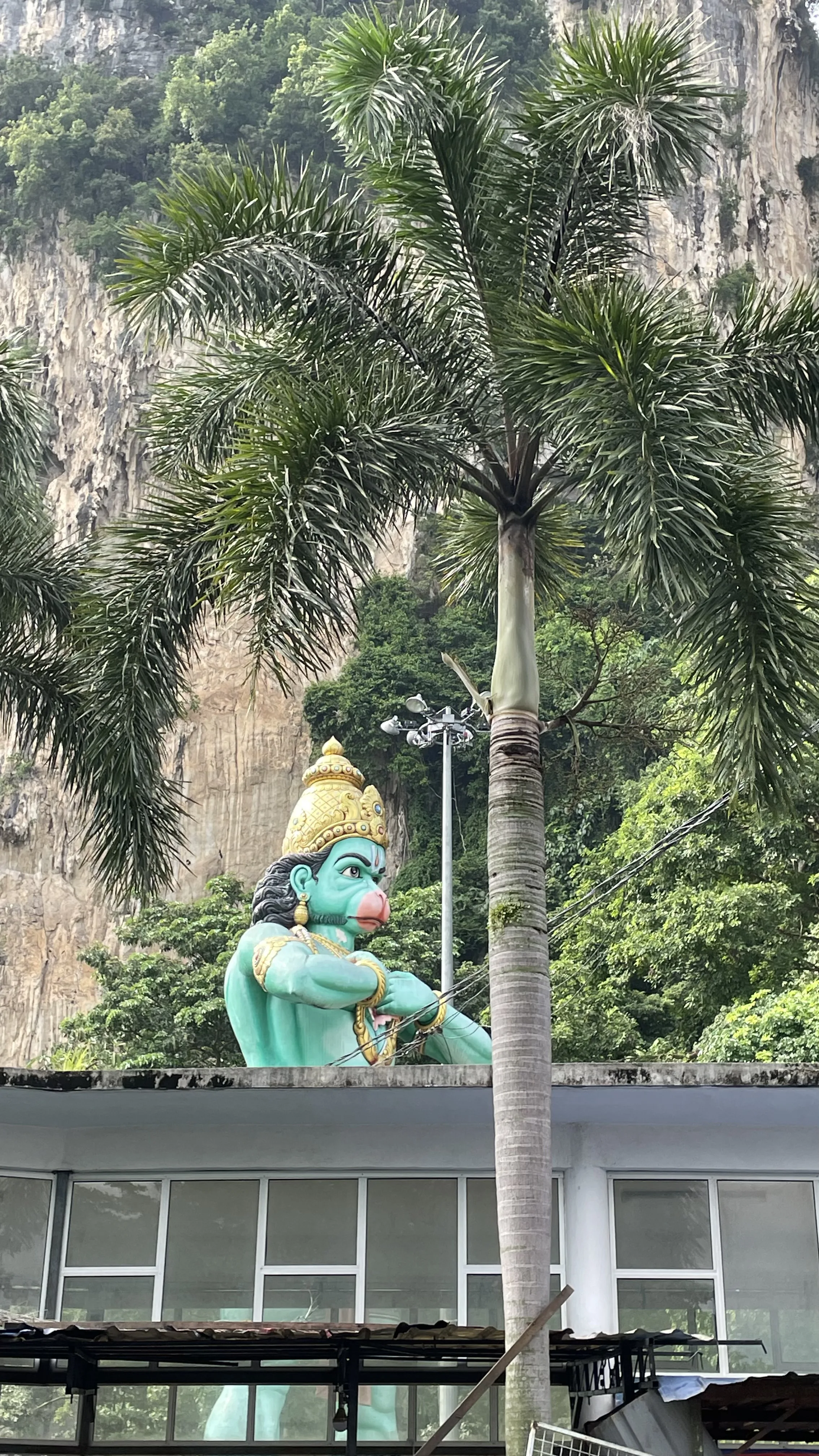 Estátua verde e dourada do deus Hanuman em meio à vegetação e paredão rochoso em Batu Caves, na Malásia.