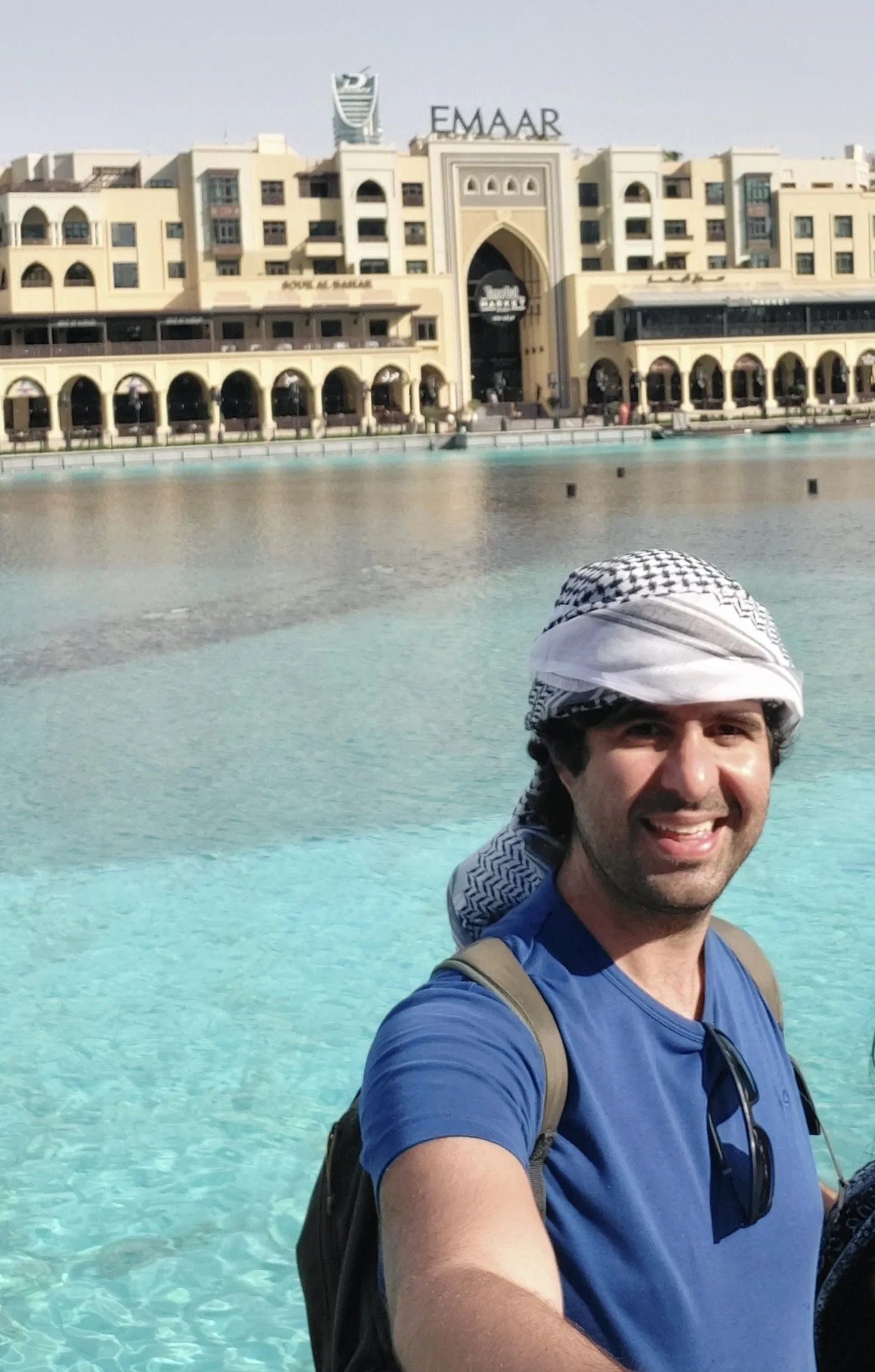 Homem em primeiro plano usando lenço keffiyeh na cabeça e camiseta azul, sorrindo de leve com a grande piscina externa e a fachada clássica do complexo Emaar e do Souk Al Bahar ao fundo em Dubai.