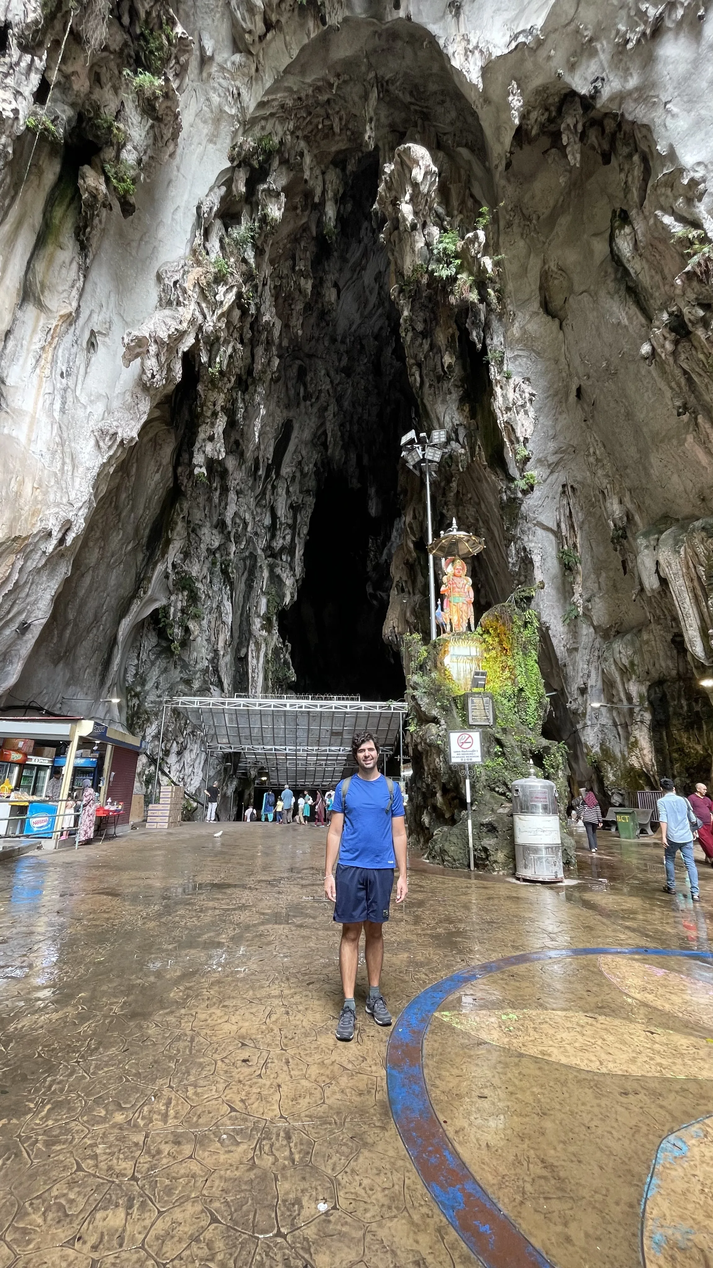 Um homem em pé no pátio molhado diante da imensa abertura rochosa e estalactites da entrada das cavernas de Batu Caves, na Malásia.