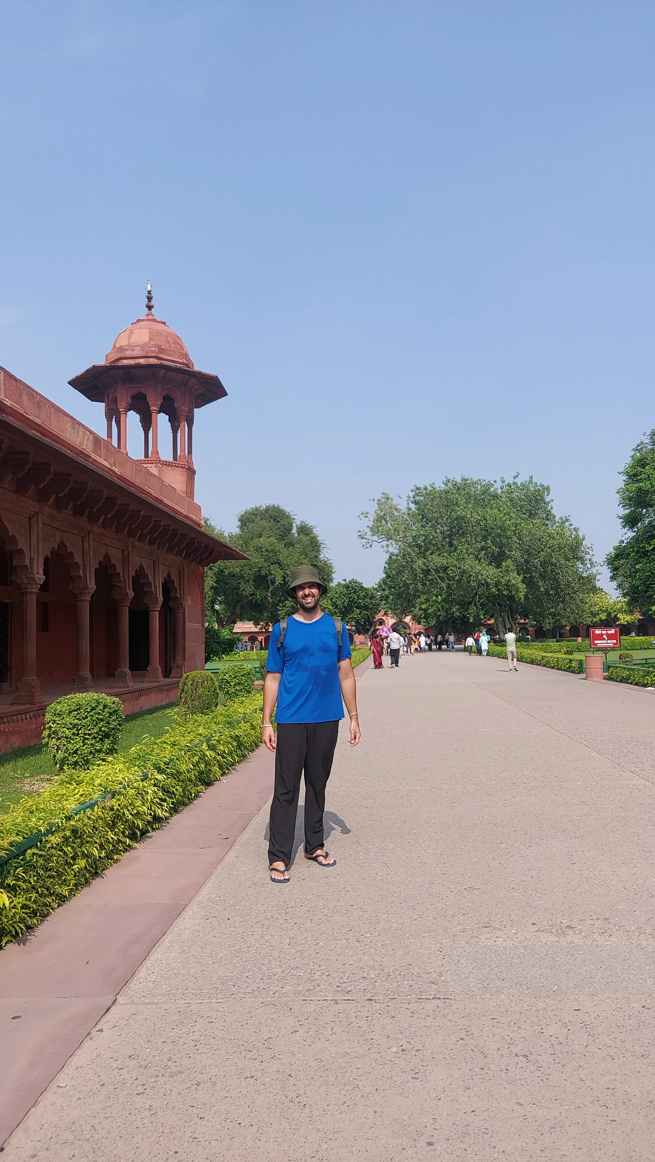 Caminho de lajes de pedra vermelha cercado por gramados verdes e arbustos podados que leva em direção ao Taj Mahal, com pedestres caminhando e o monumento de mármore branco ao fundo sob céu azul.