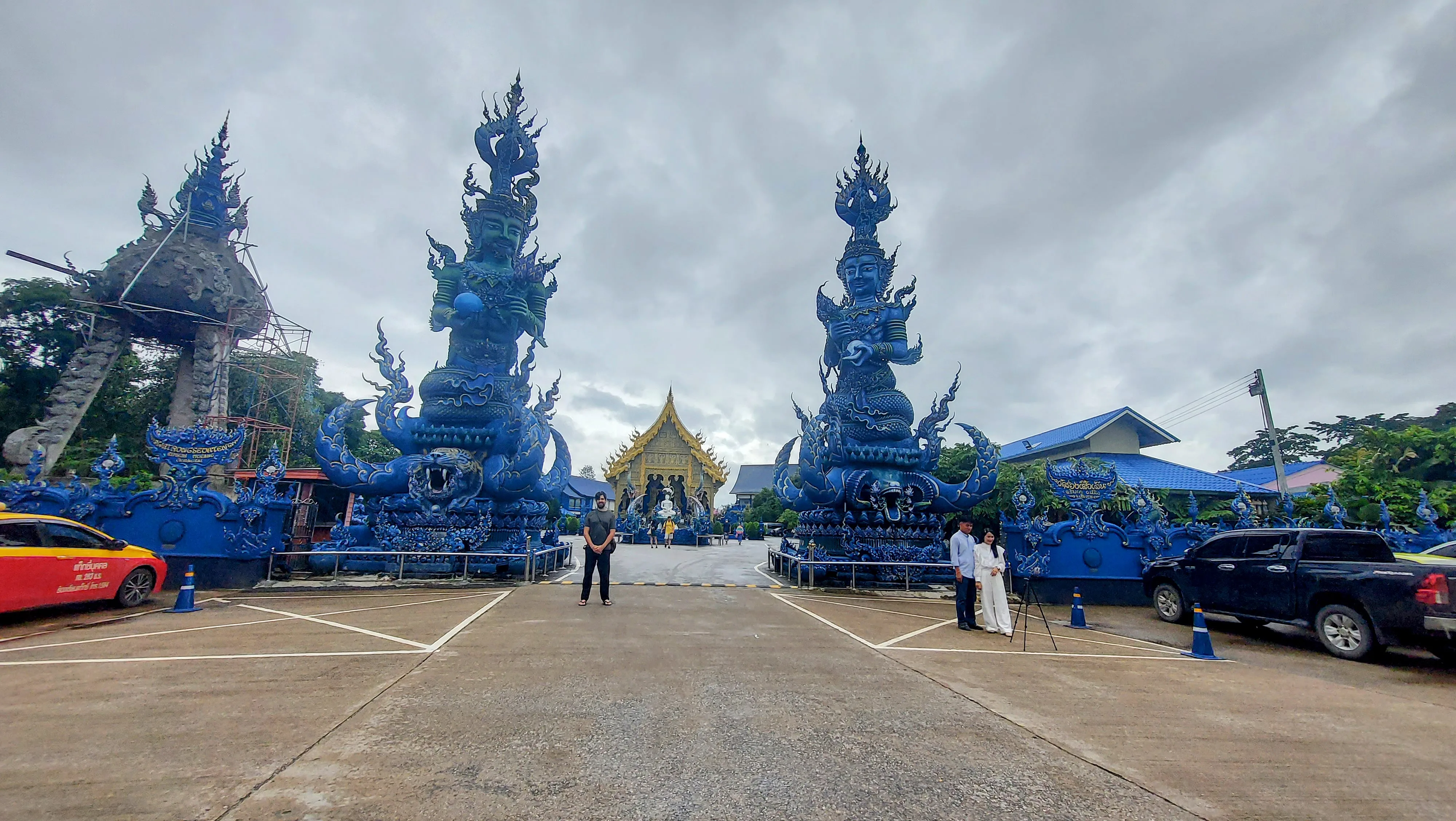 Duas grandes esculturas de guardiões azuis na entrada do Templo Azul, com a fachada principal ao fundo sob um céu nublado em Chiang Rai.