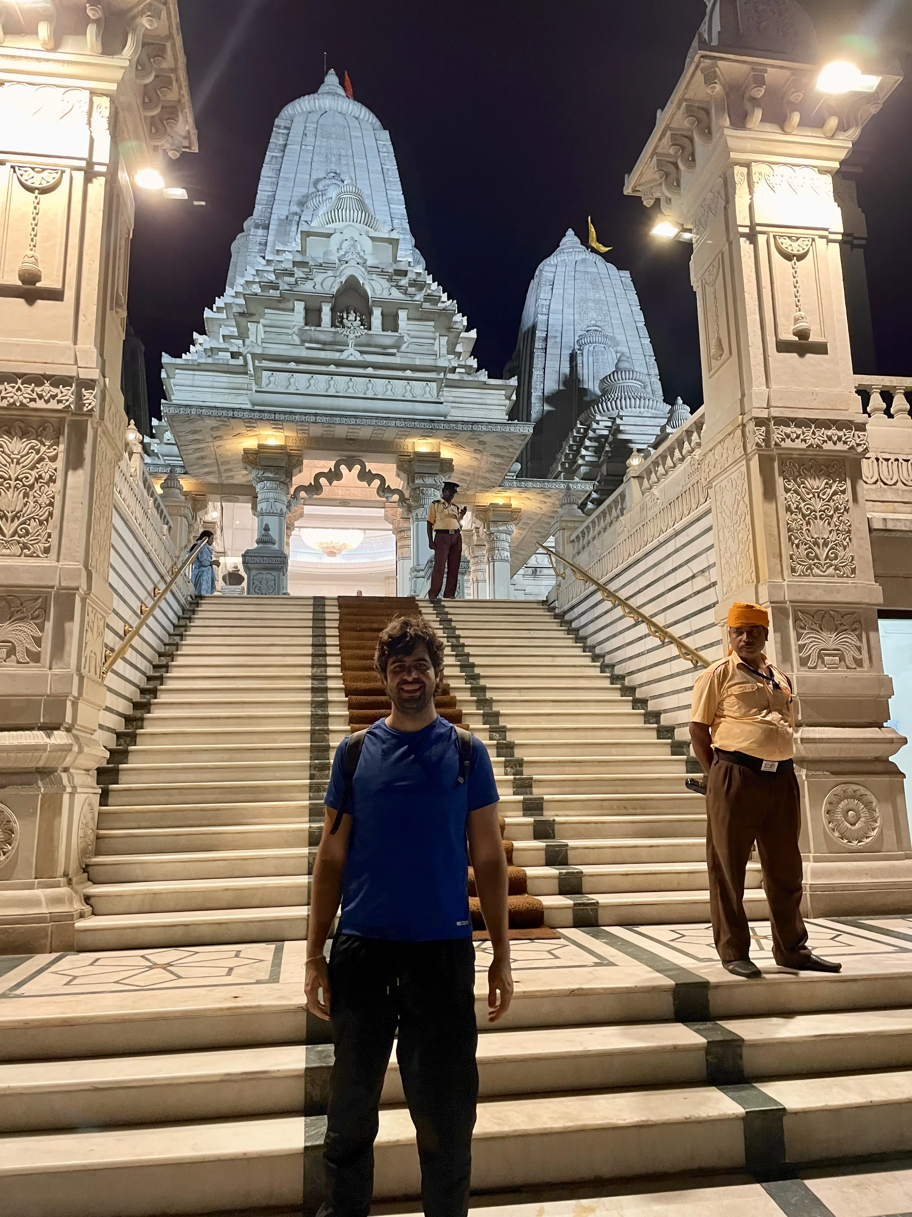 Visitante em pé na base de uma ampla escadaria de mármore branco do templo Birla Mandir à noite, ladeada por pilares ornamentados, com um segurança de uniforme ao lado e as torres iluminadas do templo ao fundo em Calcutá, Índia.