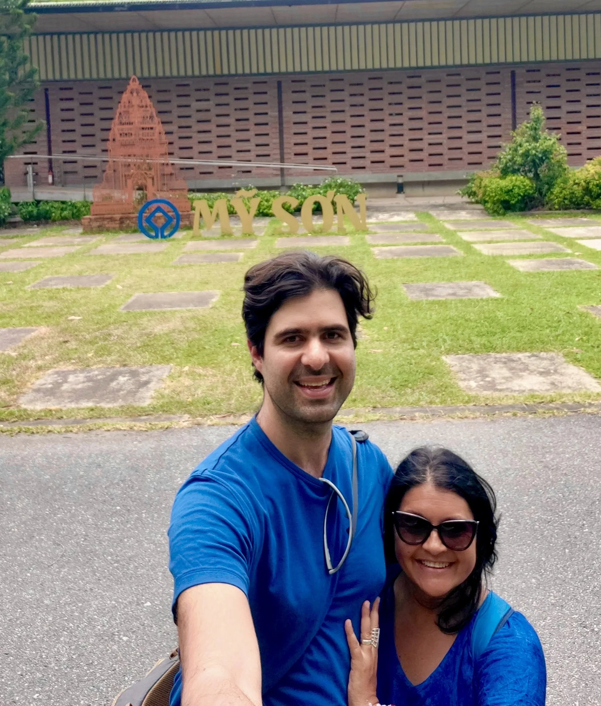 Um casal sorrindo em um selfie na frente da placa dourada 'MY SON' no local do Patrimônio Mundial, com vegetação e um edifício histórico ao fundo sob céu nublado.