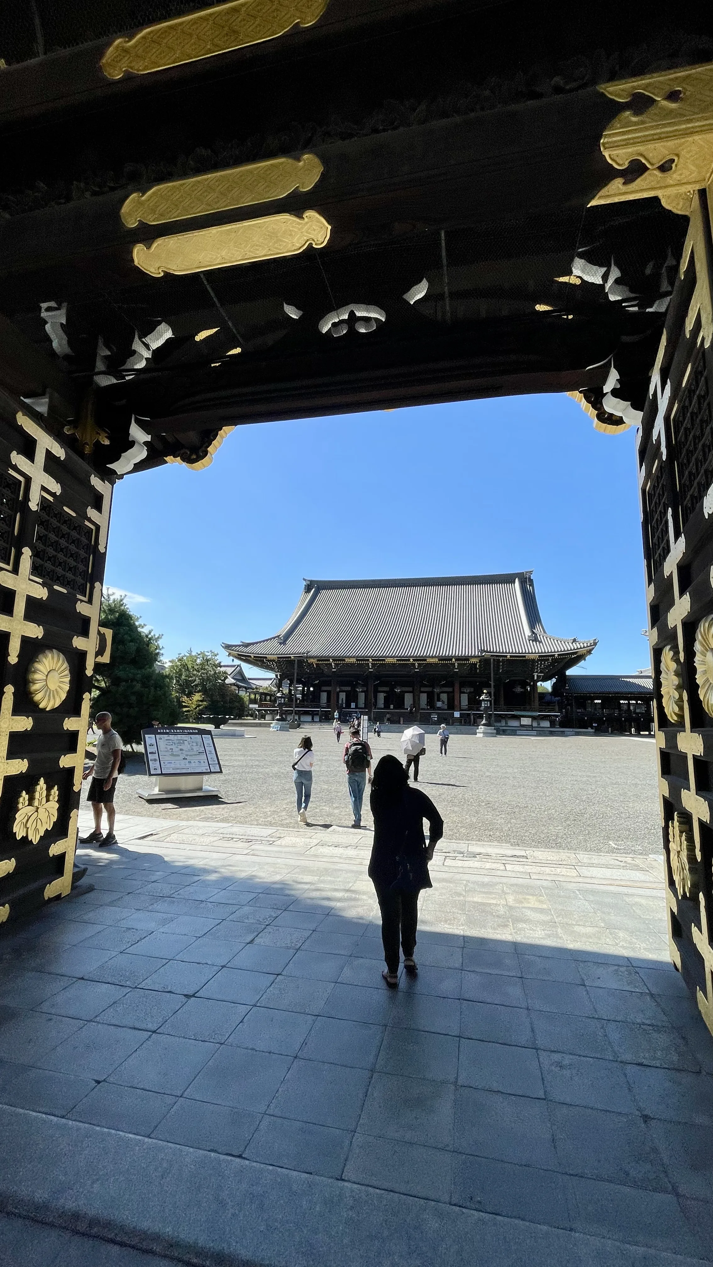 Visitantes caminhando pelo pátio de um templo em Kyoto sob um céu azul, vistos através do grandioso portão de entrada de madeira decorado com detalhes dourados