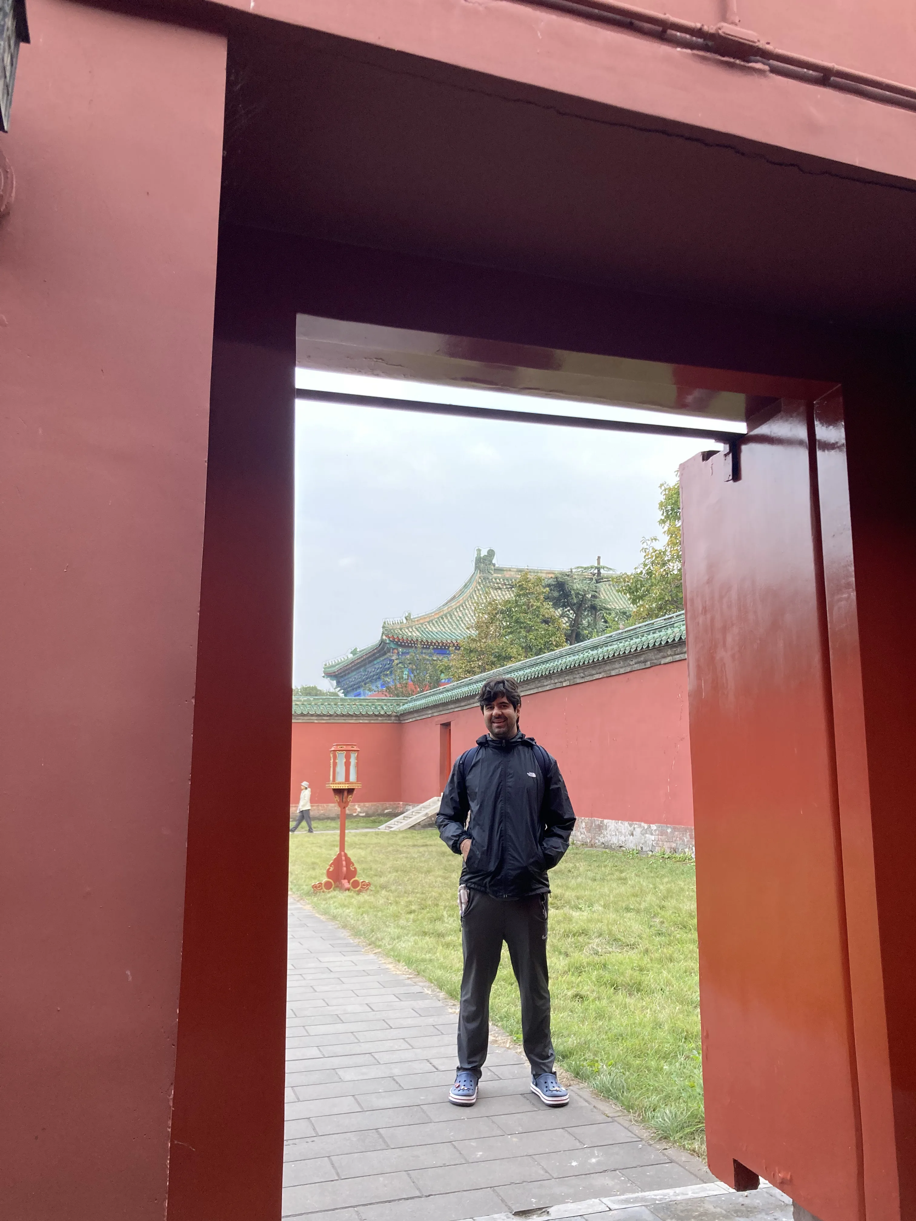 Homem sorri em pé no caminho pavimentado em frente à muralha vermelha do Templo da Abstinência em Pequim.