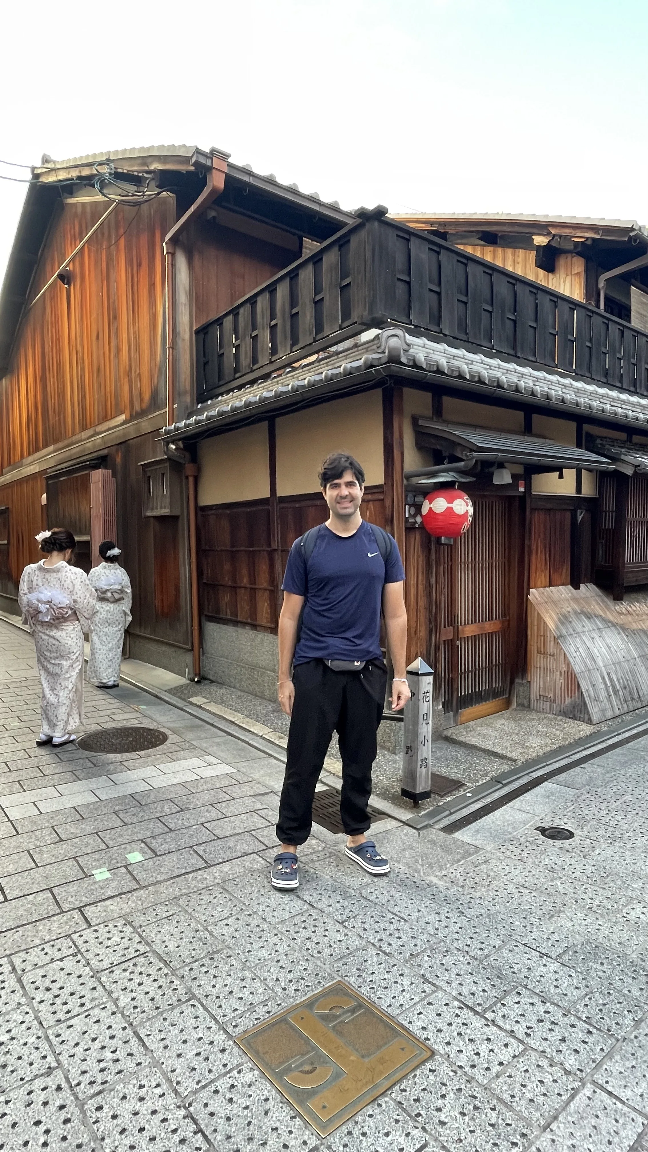 Turista posando na rua de paralelepípedos do distrito de Gion, com edifícios tradicionais de madeira e mulheres de quimono ao fundo