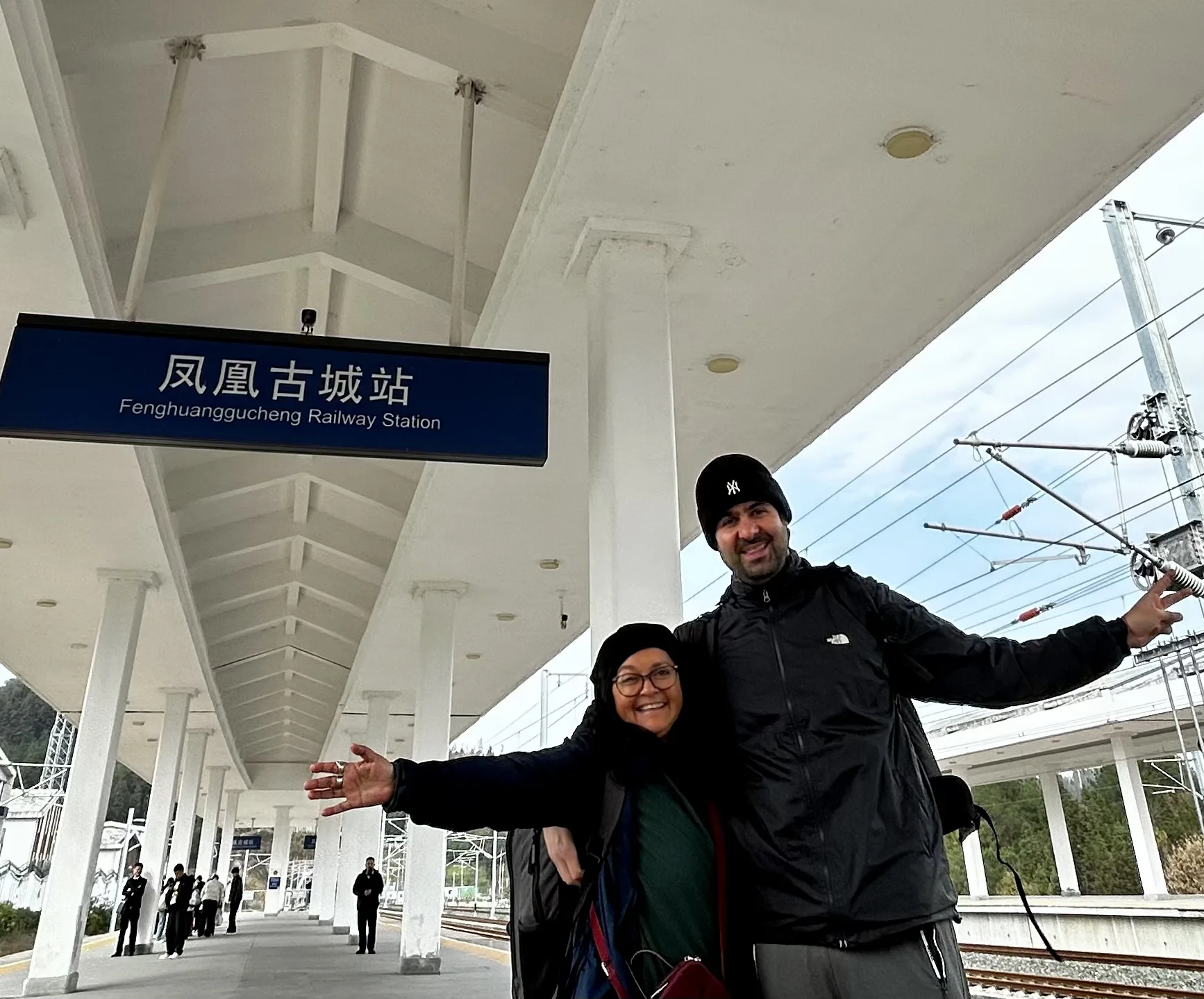 Casal posando sorridente na plataforma ao ar livre da estação ferroviária de Fenghuangzhen (FenghuangguCheng Railway Station), com os braços abertos sob a cobertura branca de pilares e os trilhos ao fundo