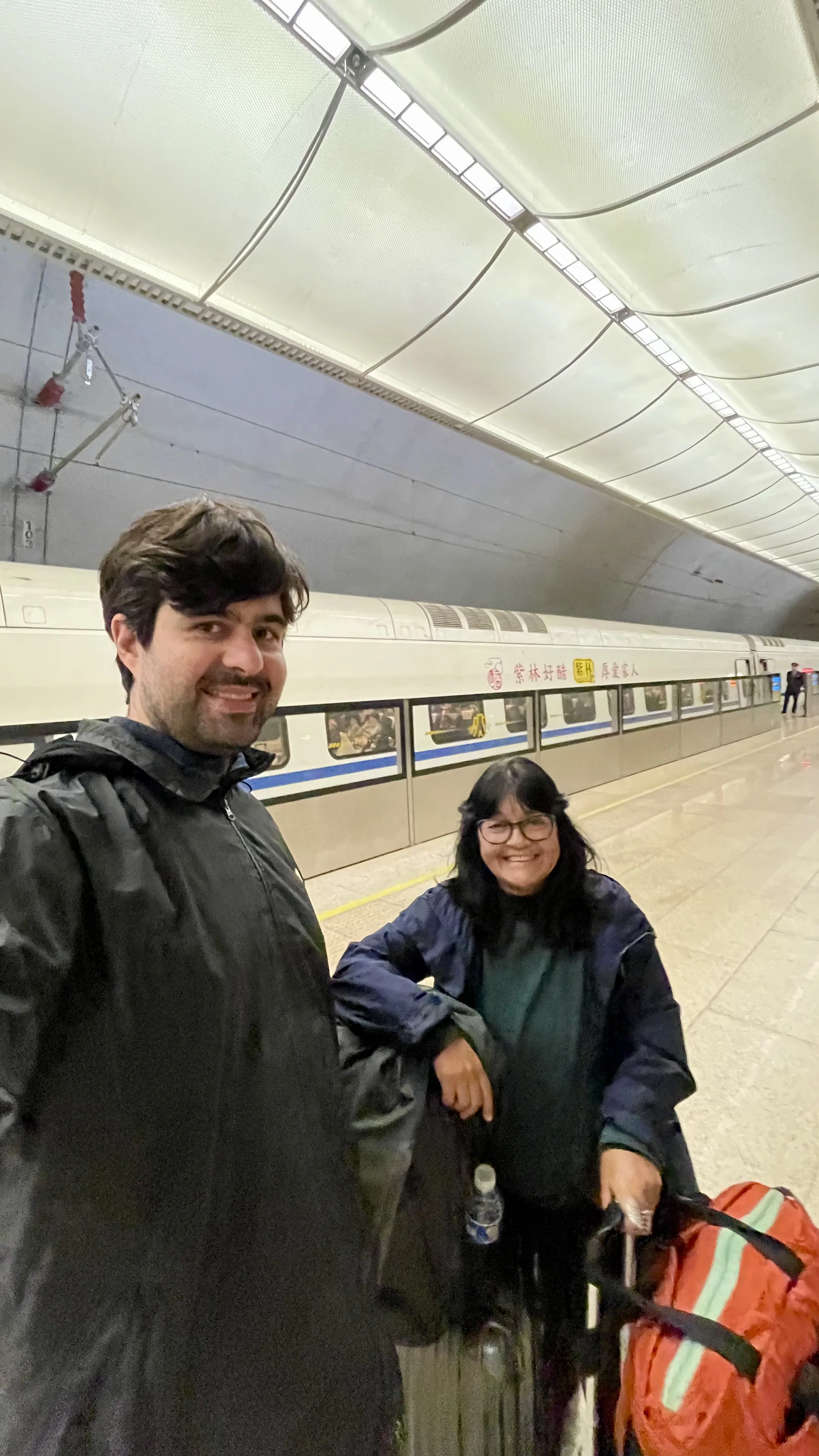 Casal de turistas sorrindo na plataforma de uma estação ferroviária moderna em Pequim, com trem bala de alta velocidade estacionado ao fundo sob teto arqueado de metal e iluminação branca.