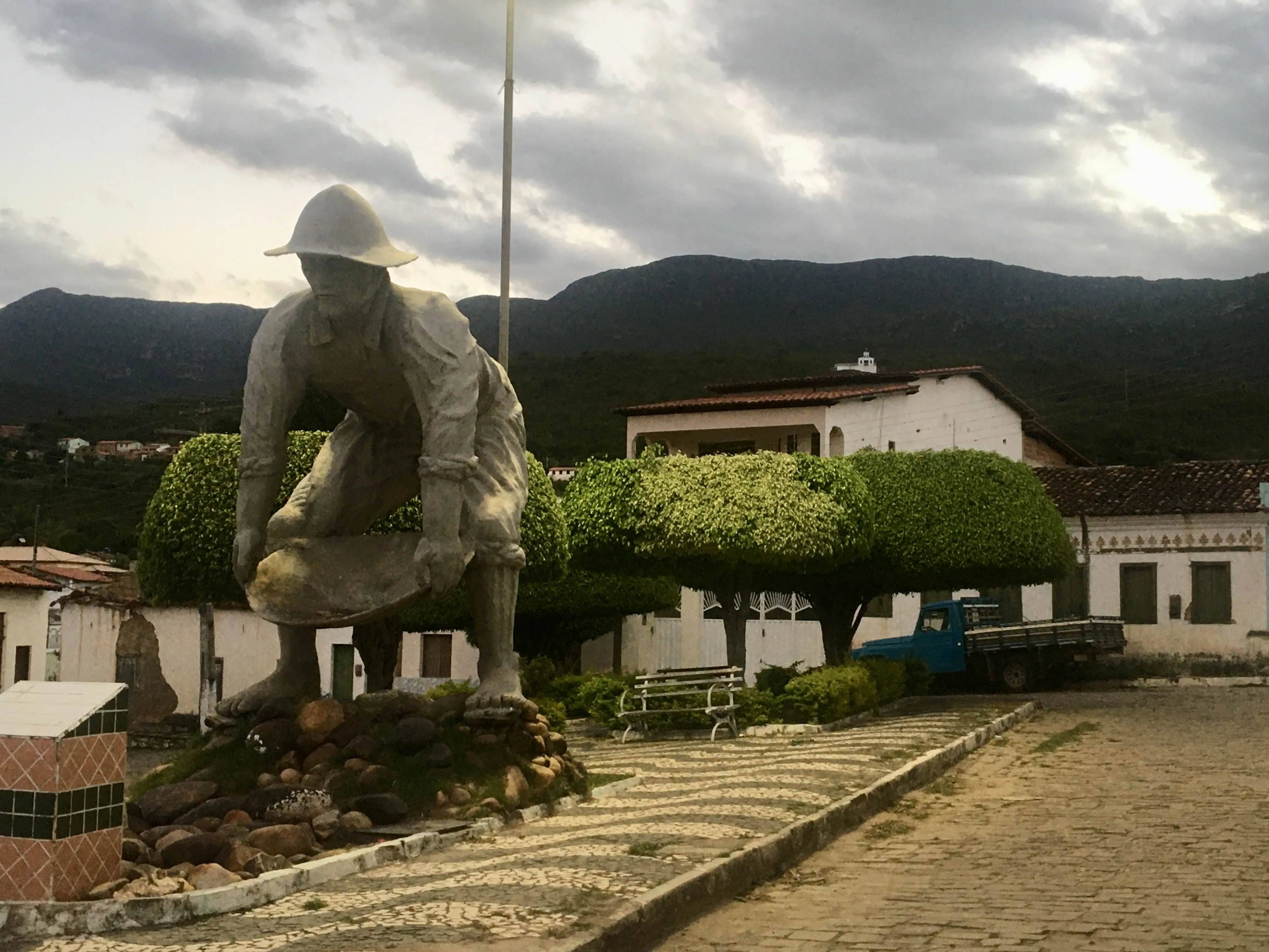 Estátua de concreto de um garimpeiro em tamanho real agachado, segurando uma bateia, no centro de uma praça de cidade pequena de Catolés. Ao fundo, montanhas sob céu nublado, casas brancas com telhados de cerâmica e árvores com copas arredondadas.