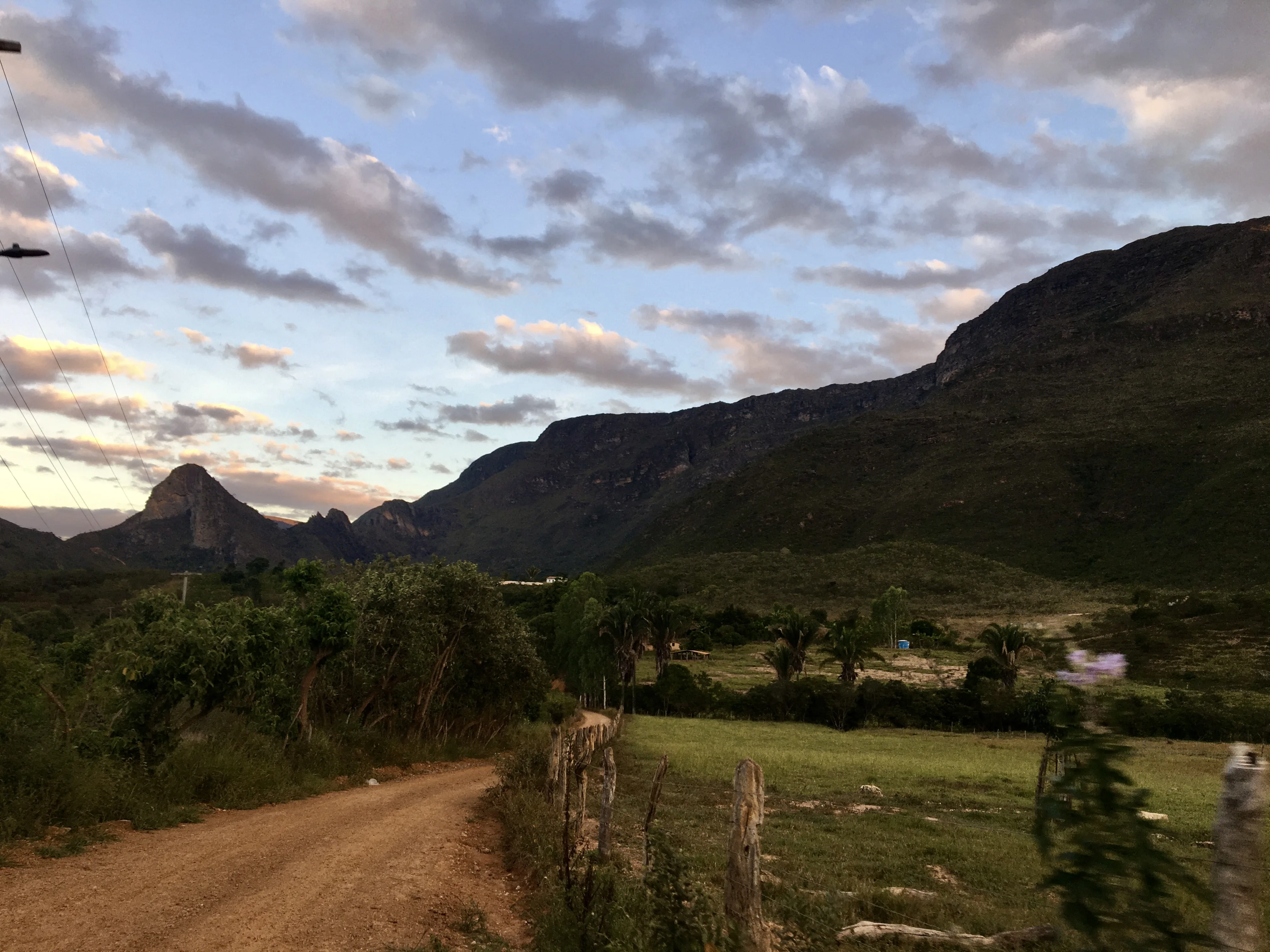 Paisagem rural com estrada de terra sinuosa serpenteando entre campos verdes e uma cerca de madeira rústica em primeiro plano. Ao longe, grandes montanhas escuras sob um céu dramático com nuvens cor-de-rosa e azuis no final da tarde