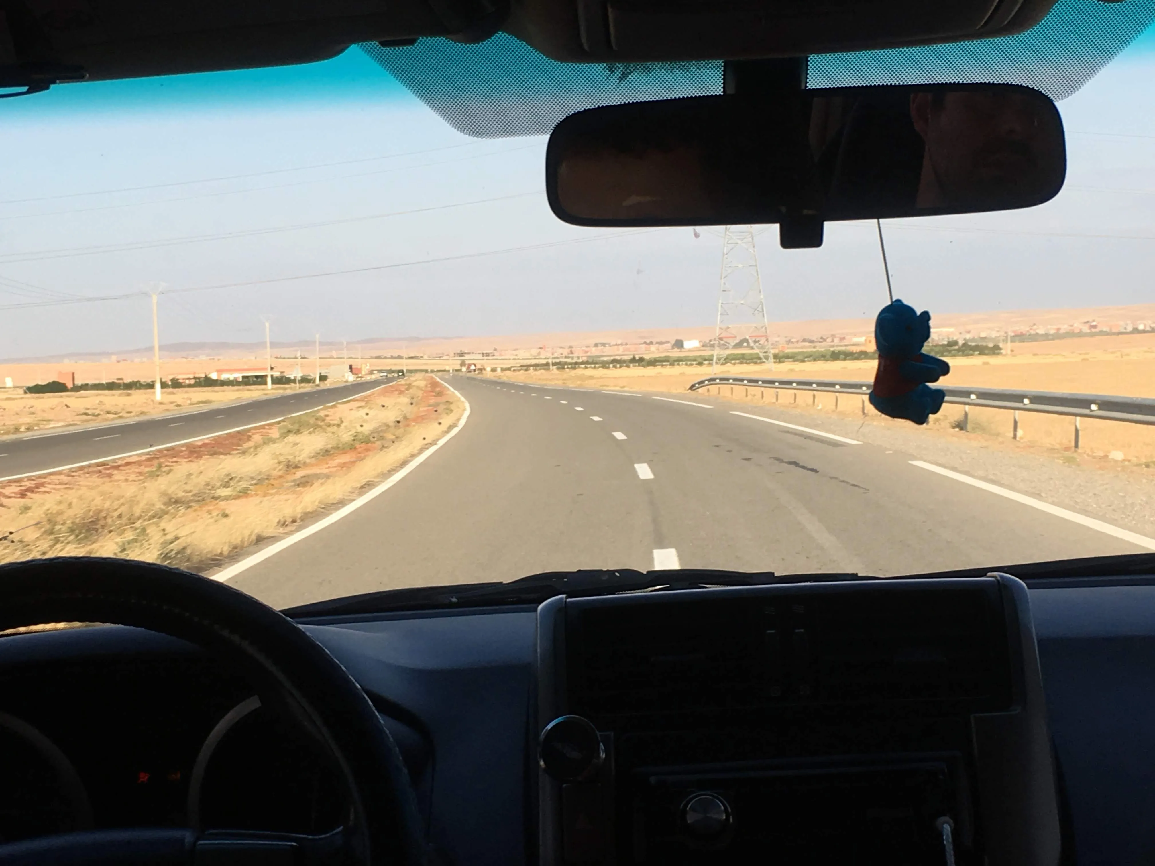 Visão de dentro de um veículo em movimento mostrando uma estrada asfaltada e vazia com curvas suaves cruzando uma paisagem árida e plana sob o céu claro na rota entre Marraquexe e Essaouira no Marrocos