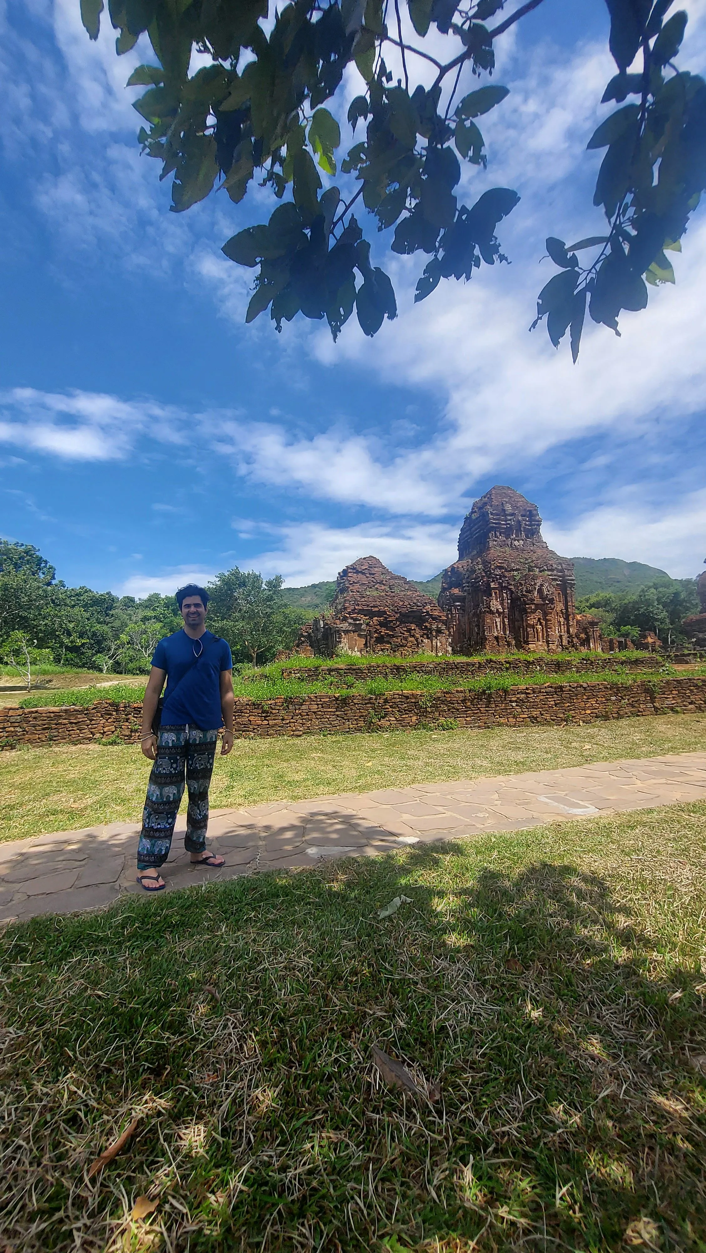Homem sorridente vestindo uma t-shirt azul e calças estampadas, em pé num caminho de pedra num relvado verde com ruínas antigas de templos de tijolos ao fundo em My Son, Vietname, sob um céu azul com nuvens emoldurado por folhas de árvores em cima.