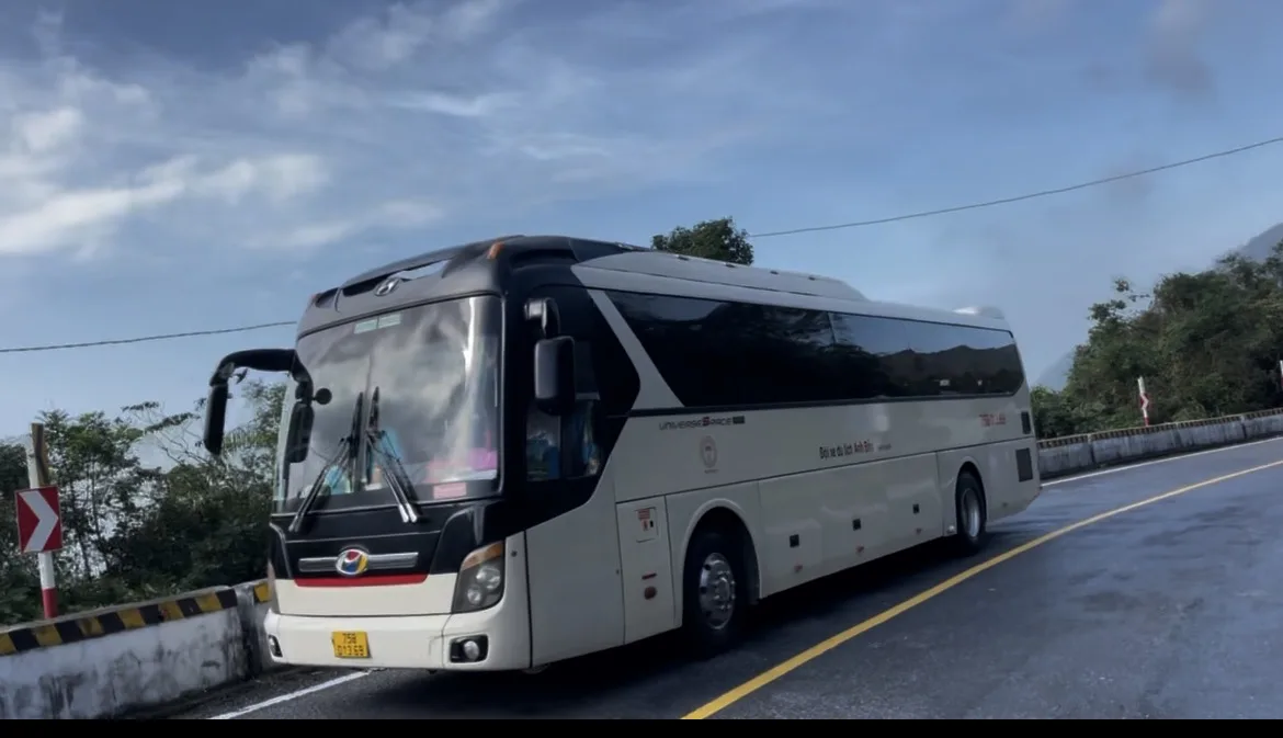 Autocarro de viagem moderno a circular numa estrada costeira e sinuosa no Vietname sob um céu azul com nuvens.