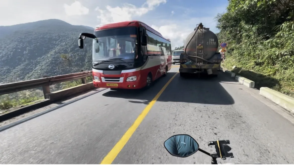 Vista a partir de uma moto em andamento numa estrada sinuosa de montanha no Vietname, com um autocarro vermelho e um camião-cisterna à frente.