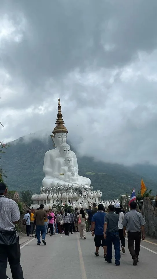 Multidão de visitantes a caminhar numa estrada em direção à imponente estátua dos cinco Budas brancos em Wat Phra That Pha Sorn Kaew sob um céu nublado.