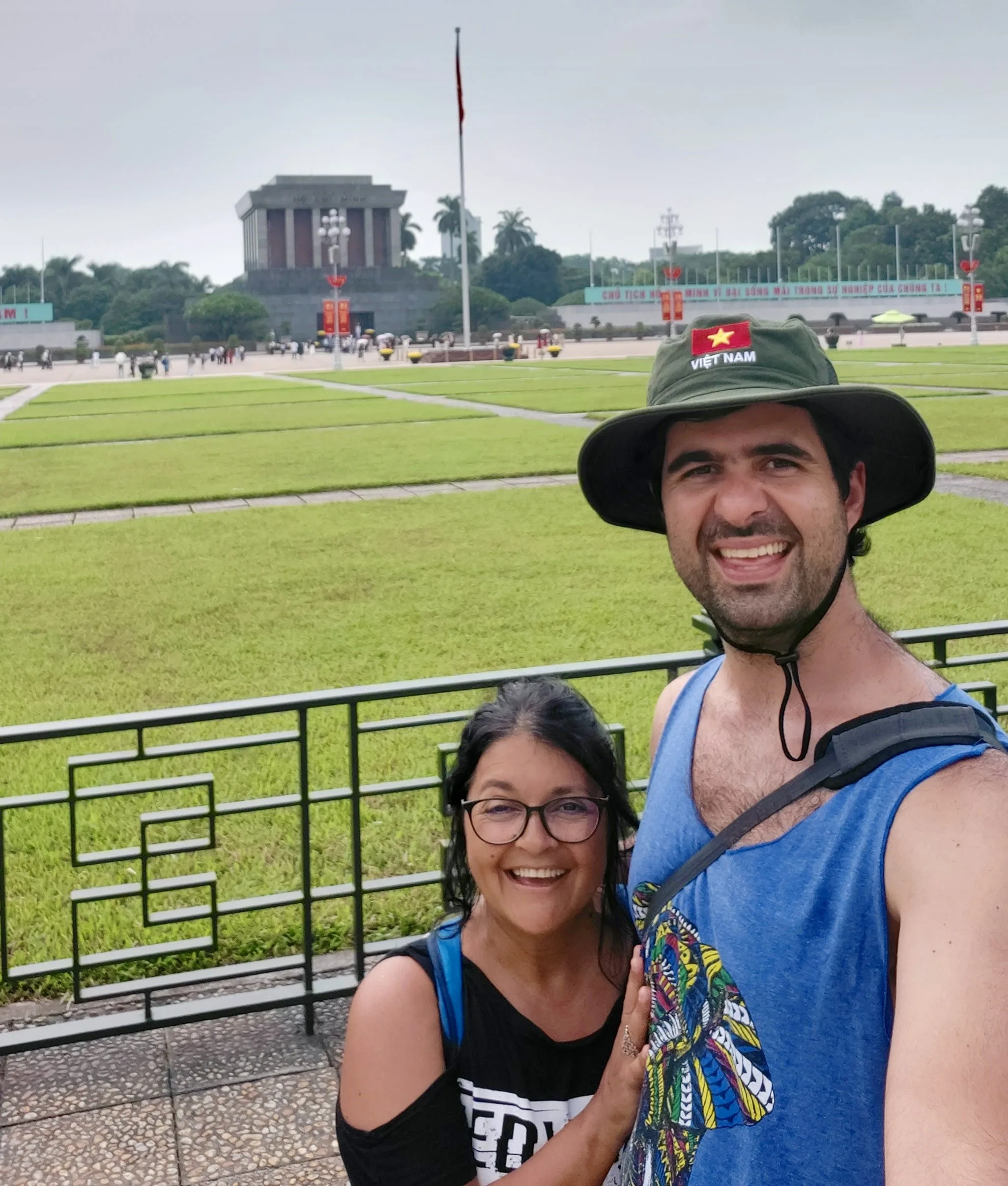 Casal sorridente em primeiro plano. Atrás deles, separa-se uma grade metálica preta e uma extensa praça pavimentada com canteiros de grama verde bem cortados. Ao fundo, destaca-se o volumoso edifício cinzento de blocos retangulares e colunas sóbrias do Mausoléu de Ho Chi Minh, sob um dia nublado.