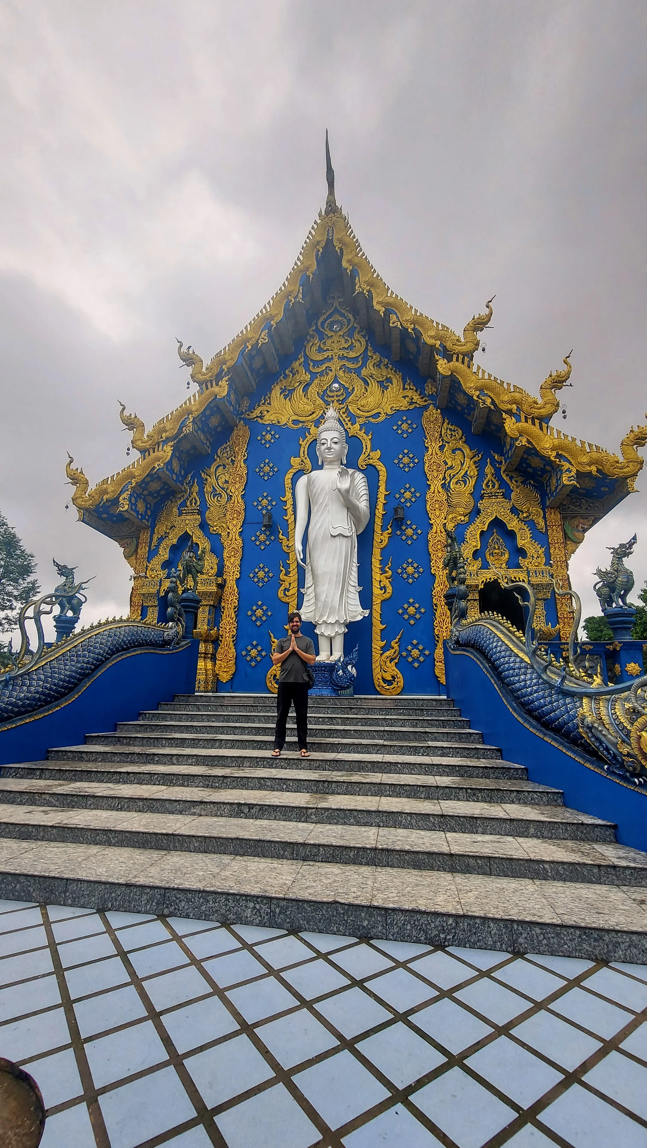 Um homem em pé com as mãos unidas em gesto de reverência na escadaria diante da fachada azul e dourada e da estátua de Buda do Templo Azul em Chiang Rai, na Tailândia.
