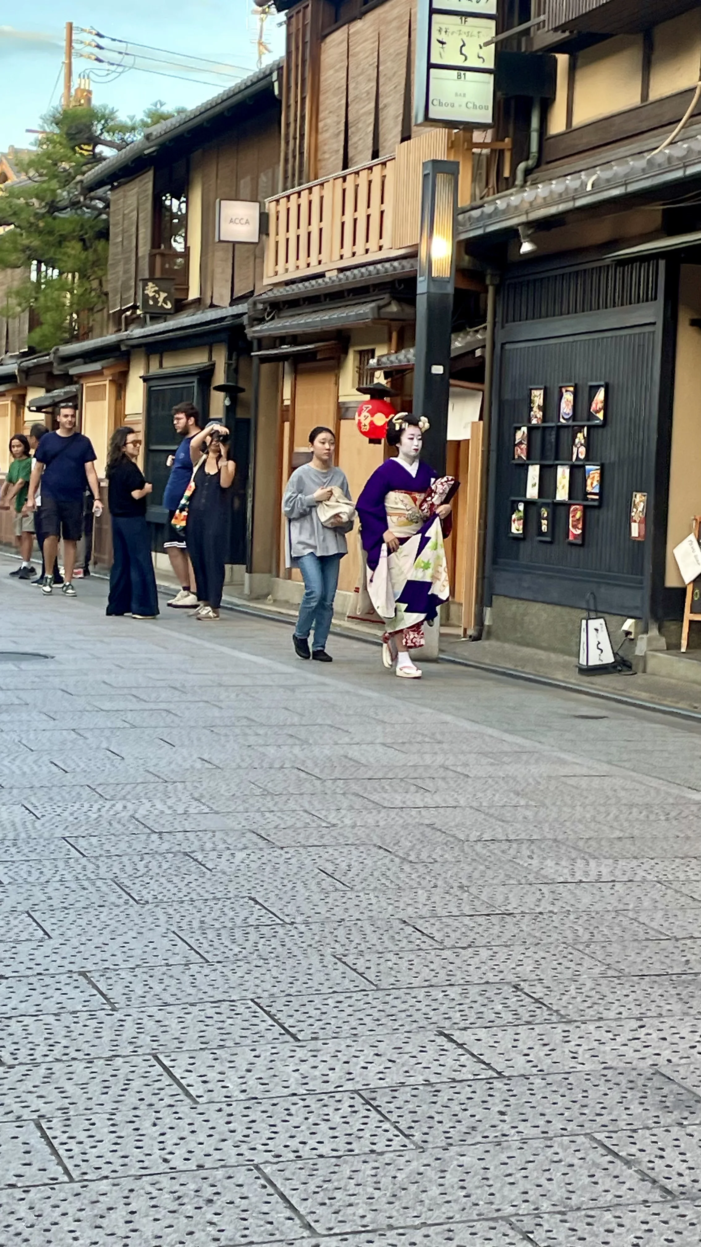 Gueixa com quimono tradicional roxo e branco caminha por uma rua de paralelepípedos em Gion, Kyoto, ladeada por edifícios de madeira e vitrines iluminadas.