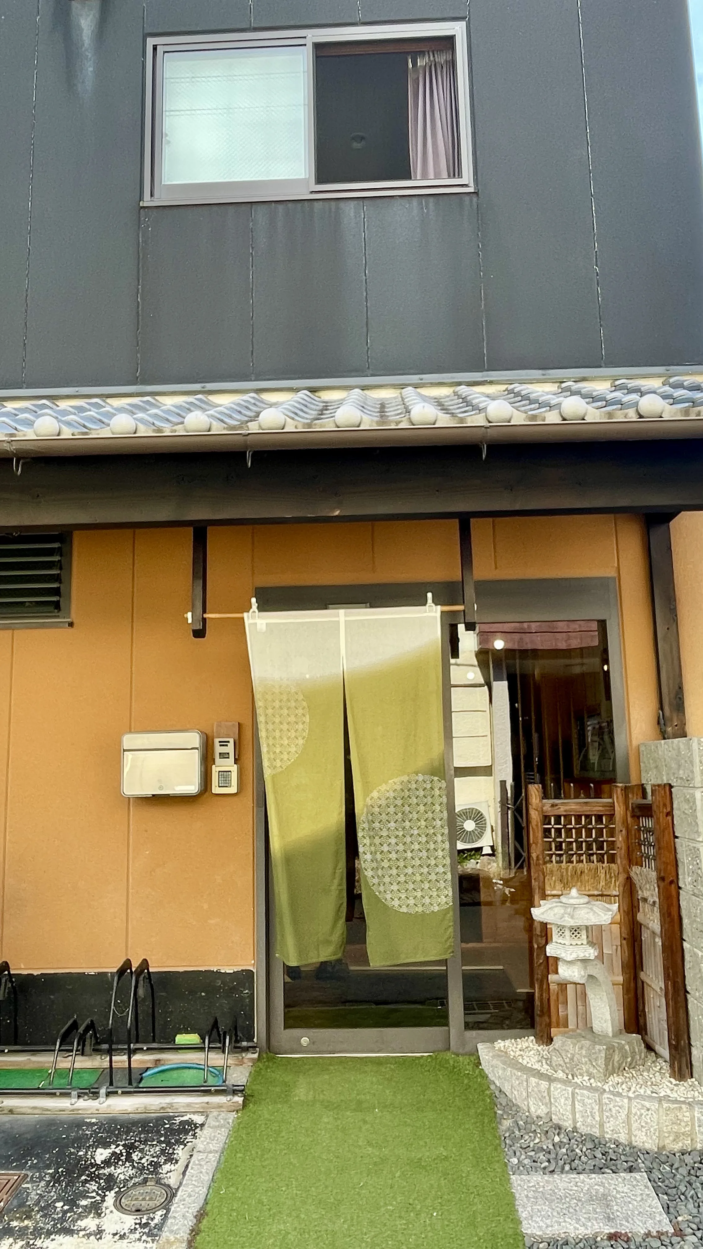 Entrada de guesthouse tradicional em Nara com cortina noren verde, tapete sintético e lanterna de pedra ao lado.