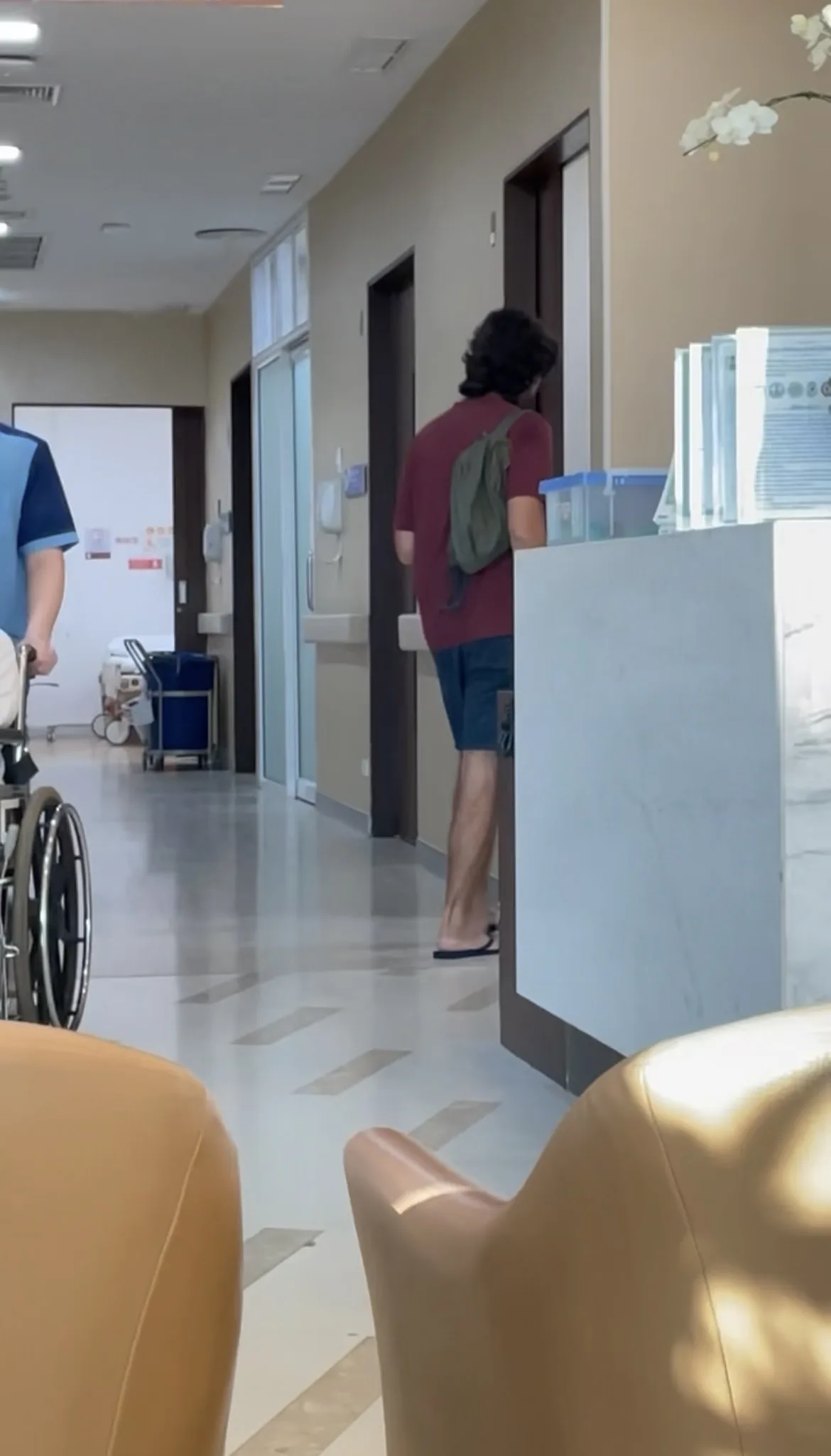 Homem de costas caminhando pelo corredor de hospital em Chiang Mai perto do balcão da recepção e de um enfermeiro com cadeira de rodas.