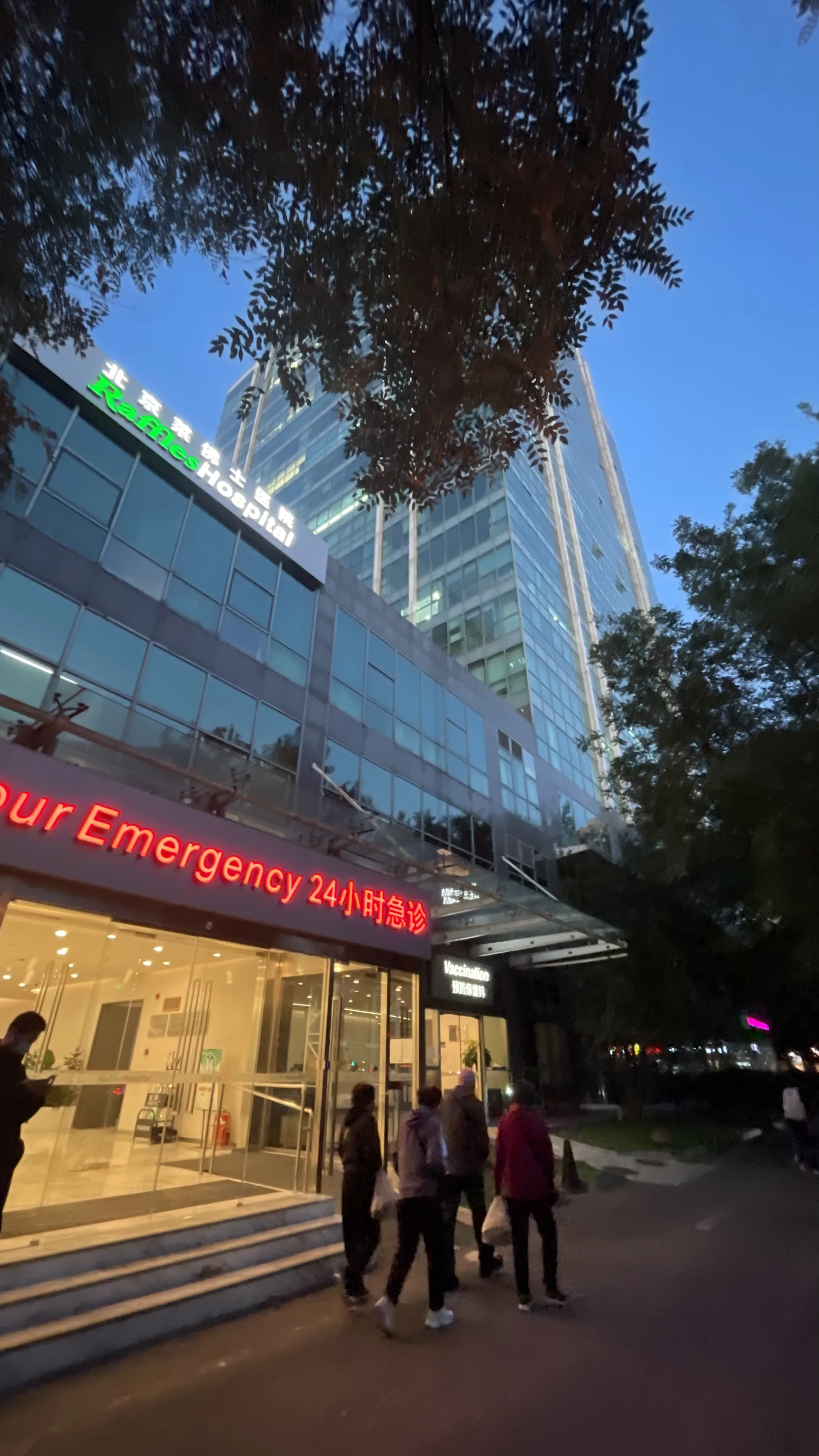 Fachada iluminada à noite do Raffles Hospital em Pequim com letreiro vermelho da emergência 24 horas e pedestres na calçada.